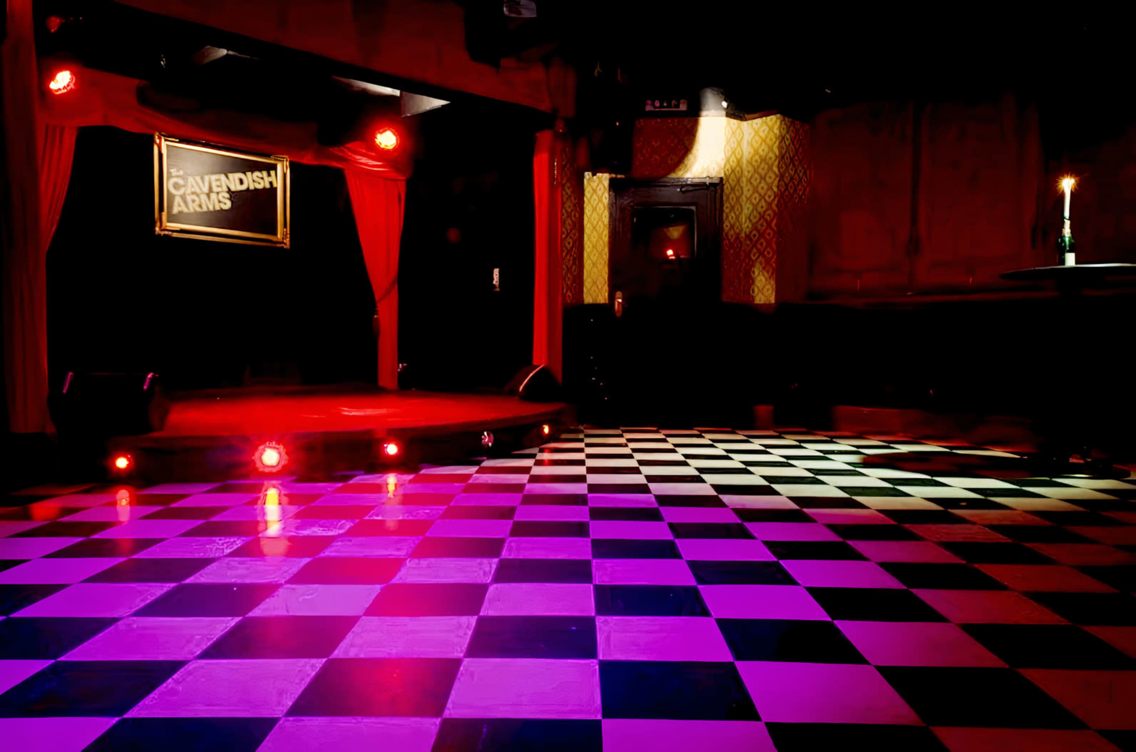 A staged area with a checkered floor and red curtains, illuminated by stage lights at the Cavendish Arms venue.
