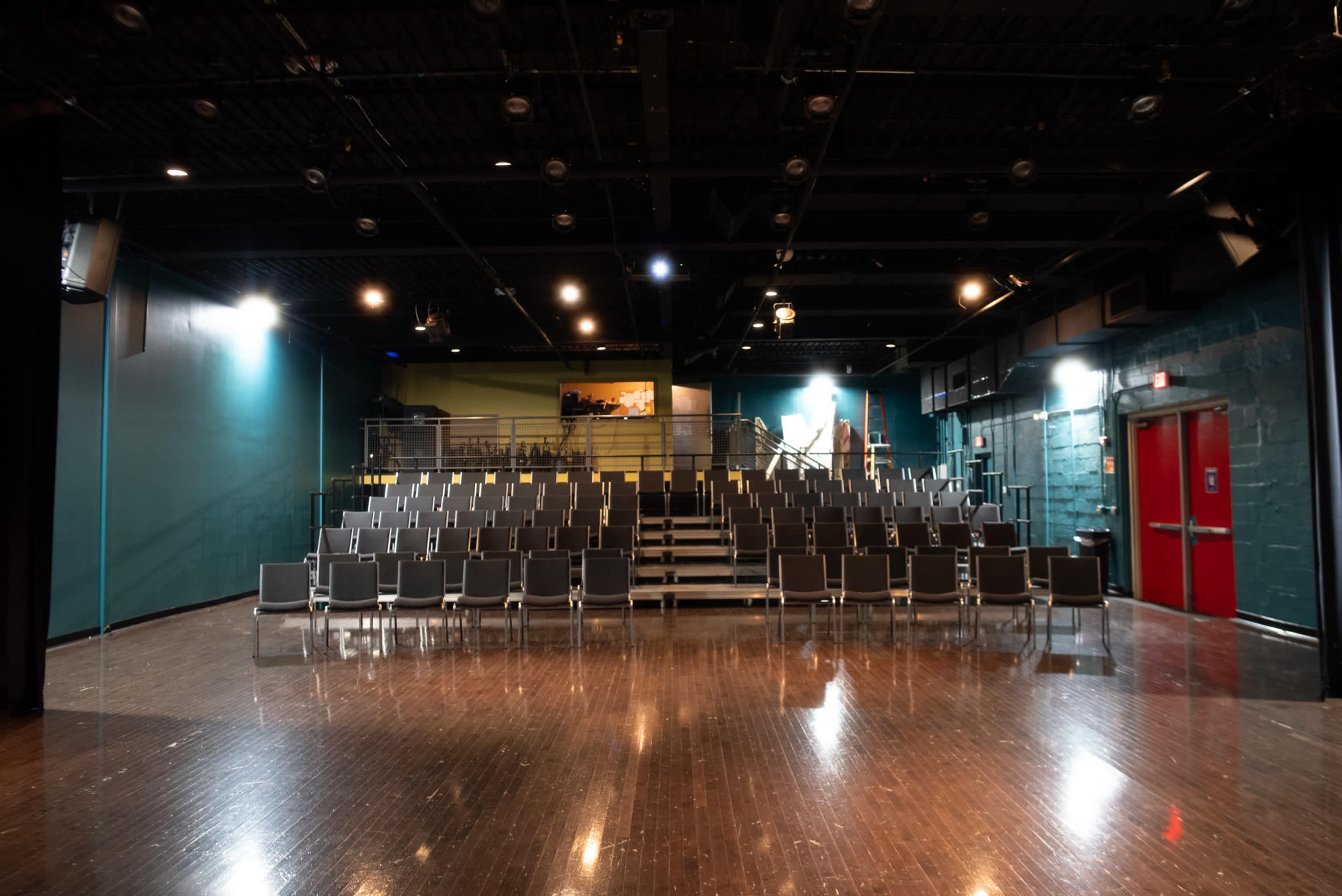 A modern theater with rows of empty chairs facing a stage area and a red door in the rear.