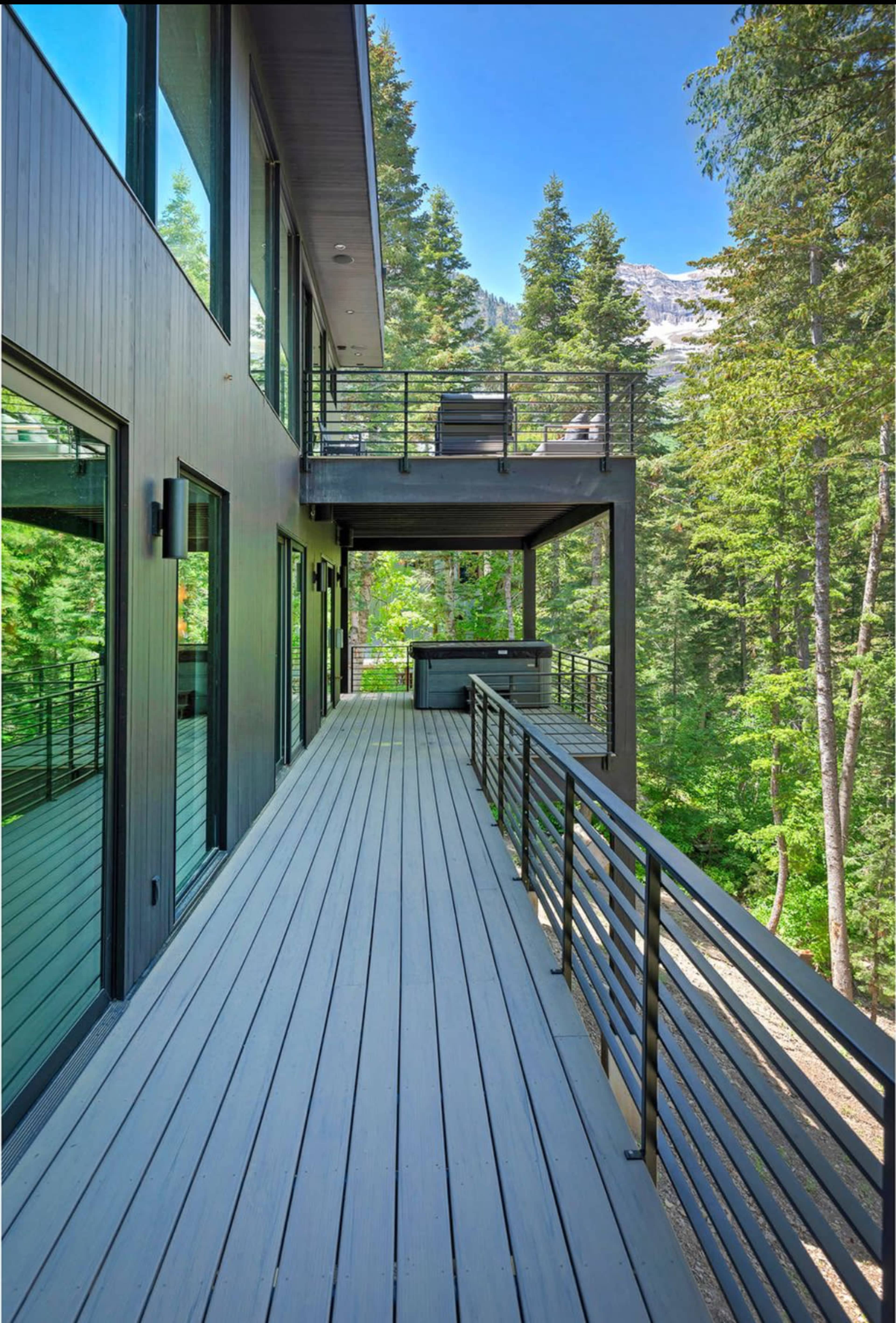 A modern building features a spacious wooden deck surrounded by tall trees and mountains in the background.