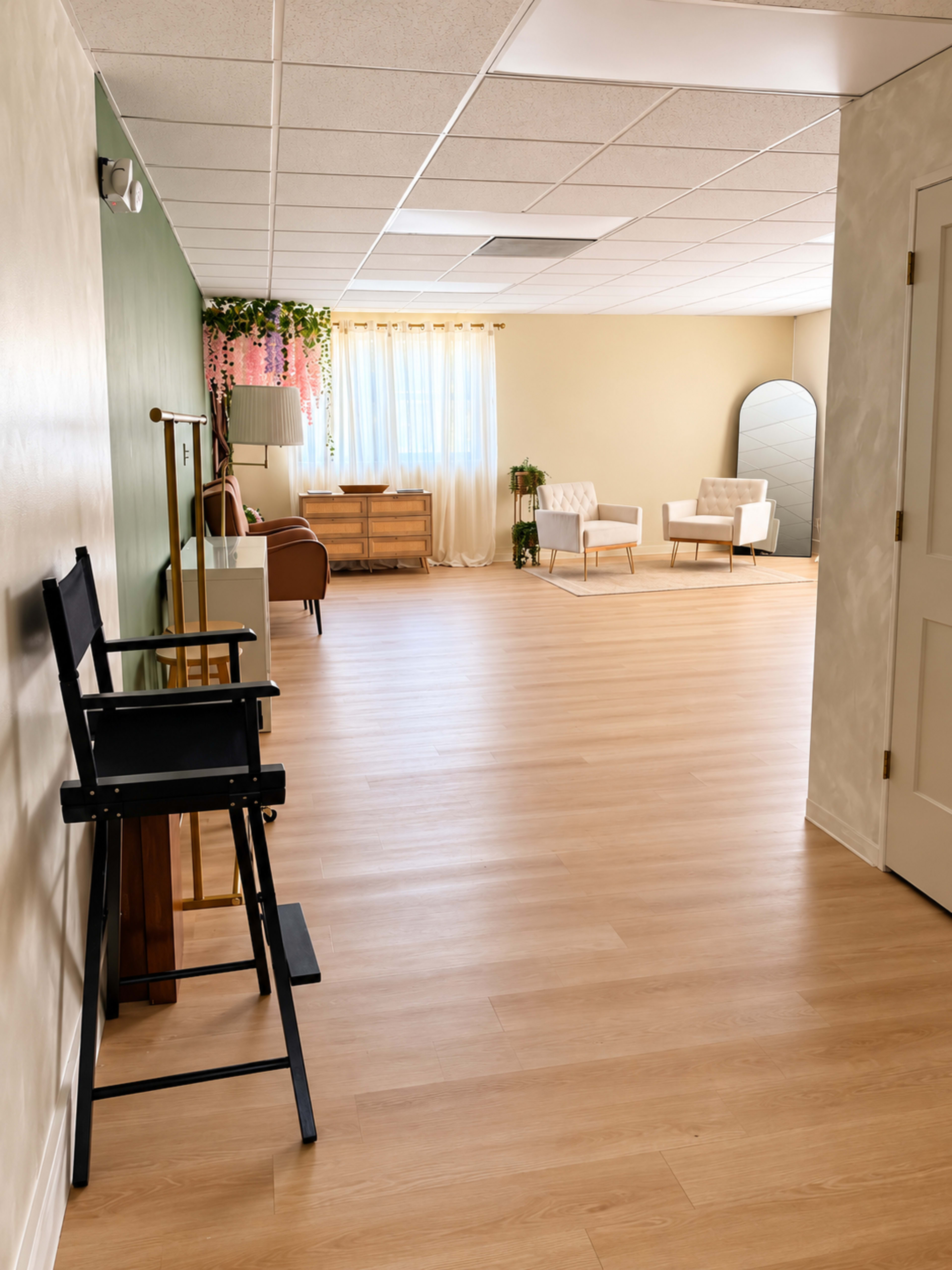 The image shows a spacious, well-lit room with a wooden floor, featuring a seating area with two chairs, a couch, and decorative plants.