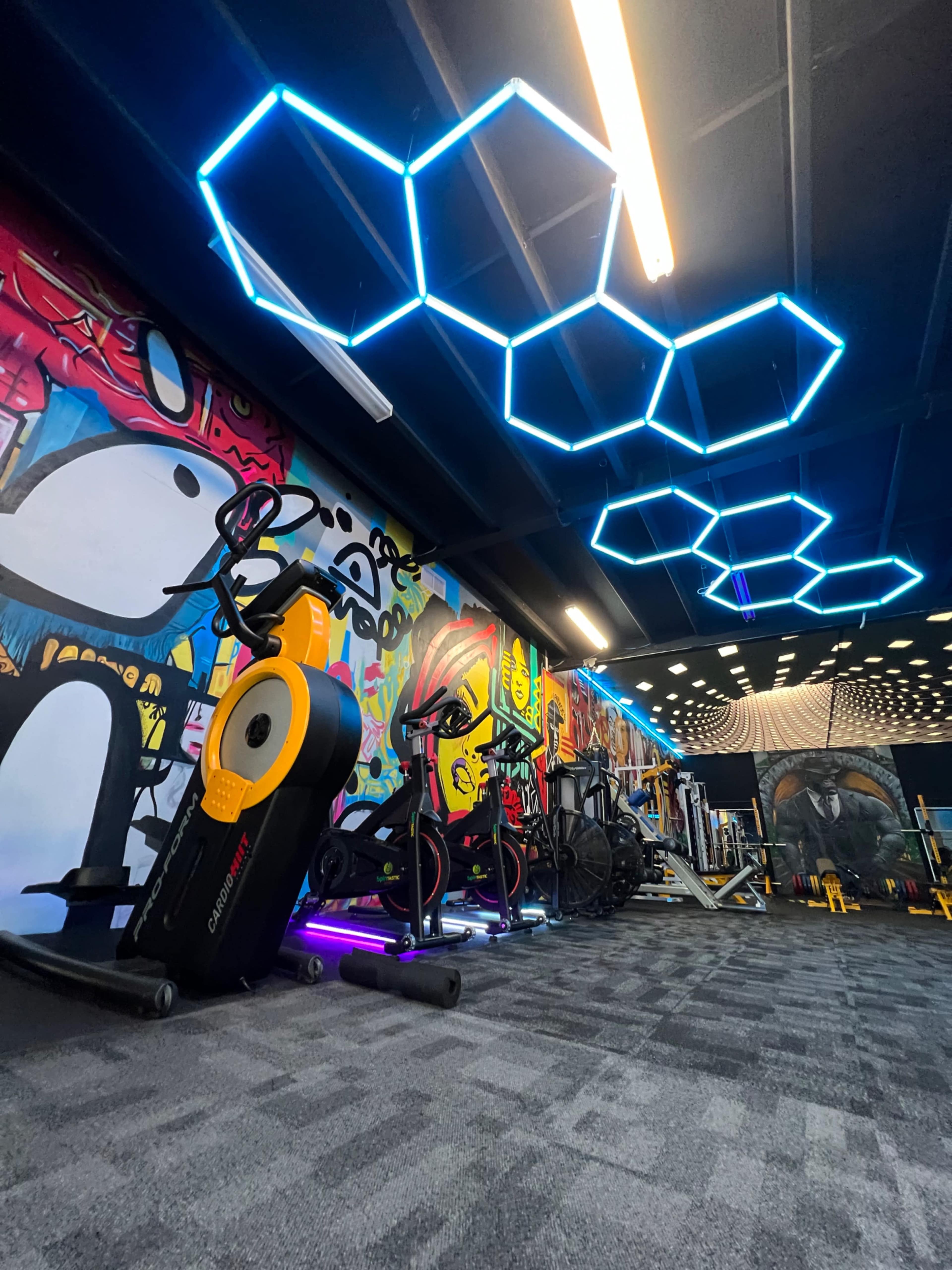 A modern gym interior featuring colorful graffiti on the walls, equipped with exercise machines and illuminated by geometric blue LED light fixtures on the ceiling.