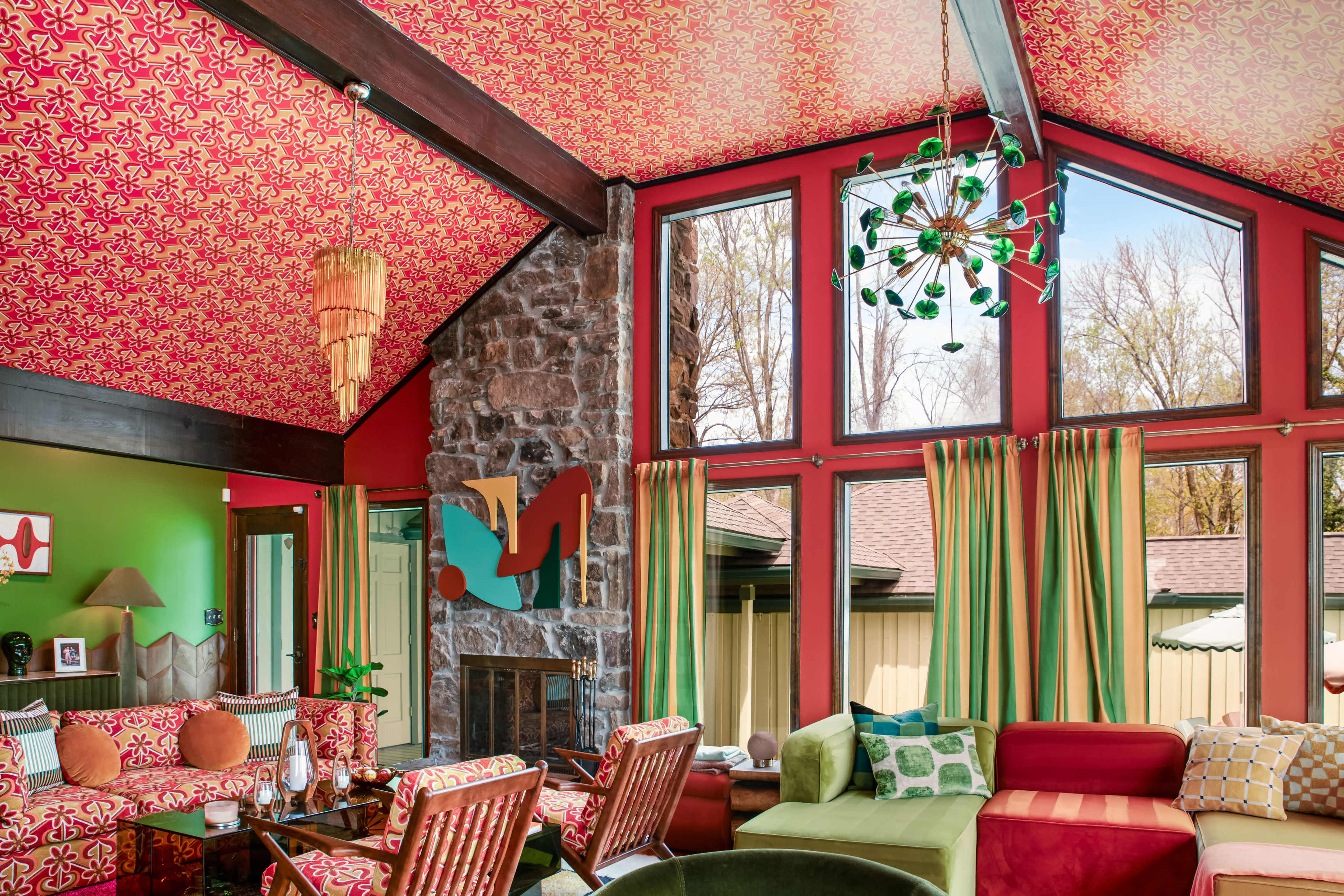 A colorful living room features a high ceiling with patterned wallpaper, large windows, a stone fireplace, and a mix of vibrant furniture arrangements.