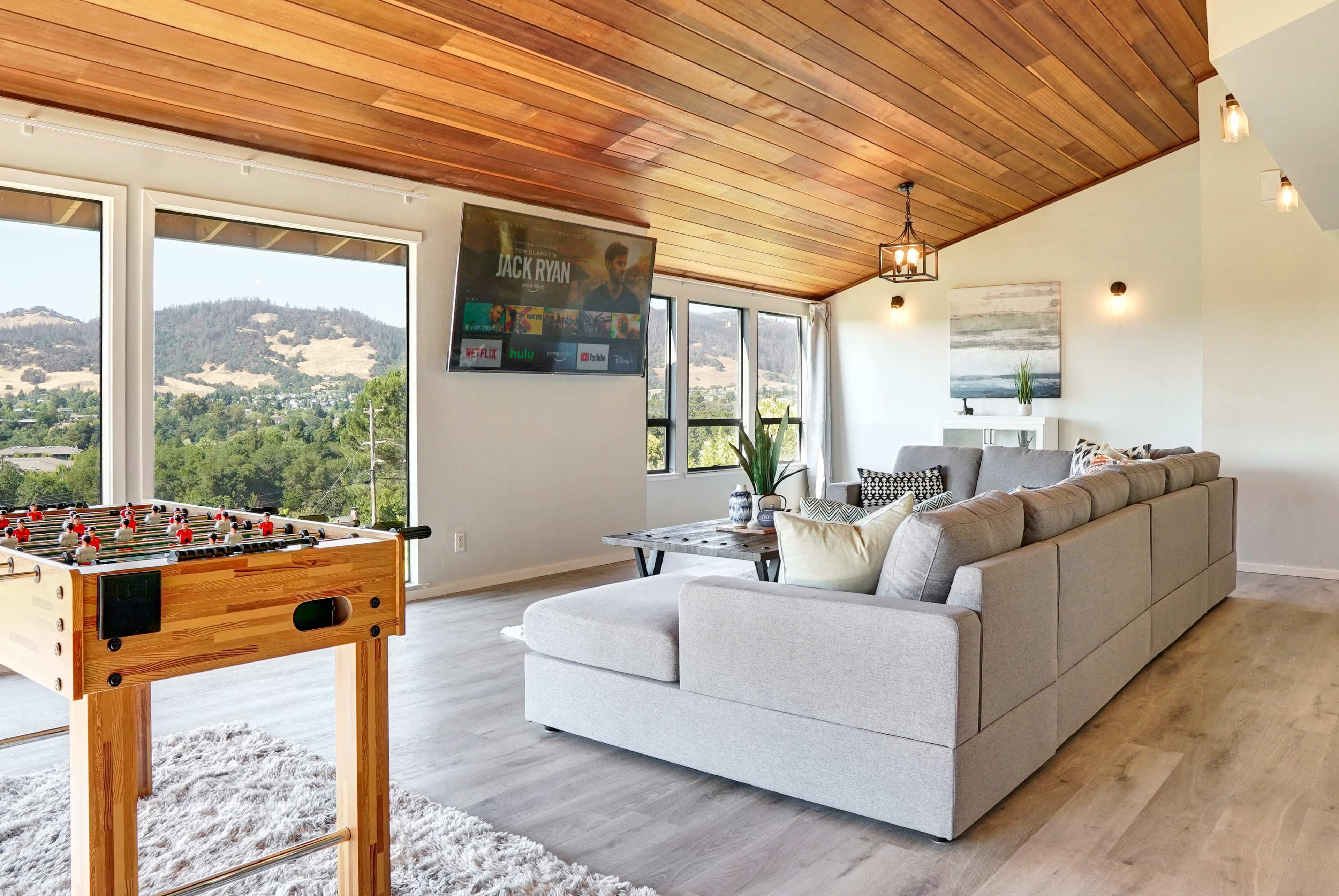 The image shows a modern living room with a large sectional sofa, a foosball table, a mounted television, and large windows overlooking a scenic landscape.