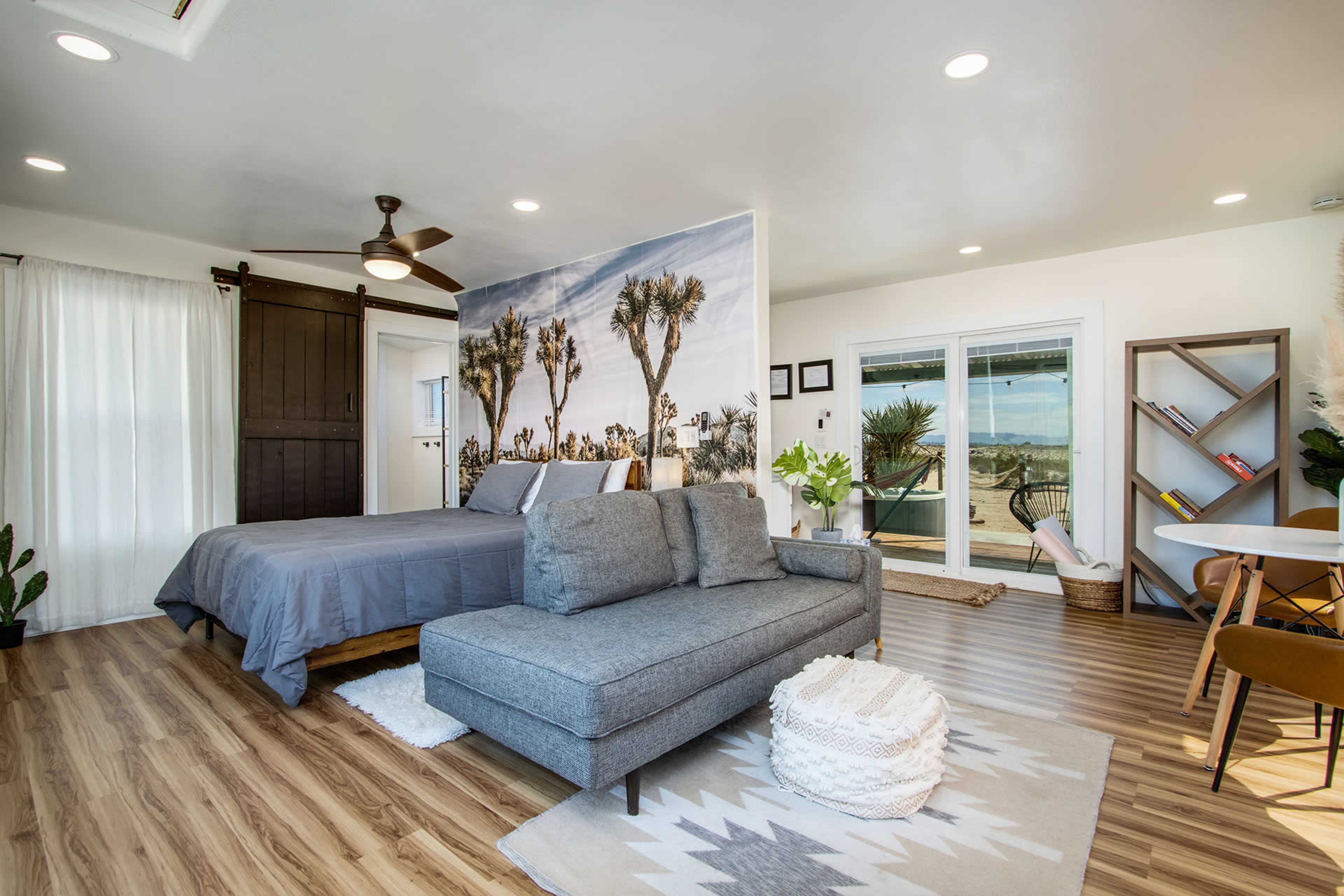 The image shows a modern bedroom with a gray bed, a sectional sofa, large windows with a view, and a mural of desert landscapes on one wall.