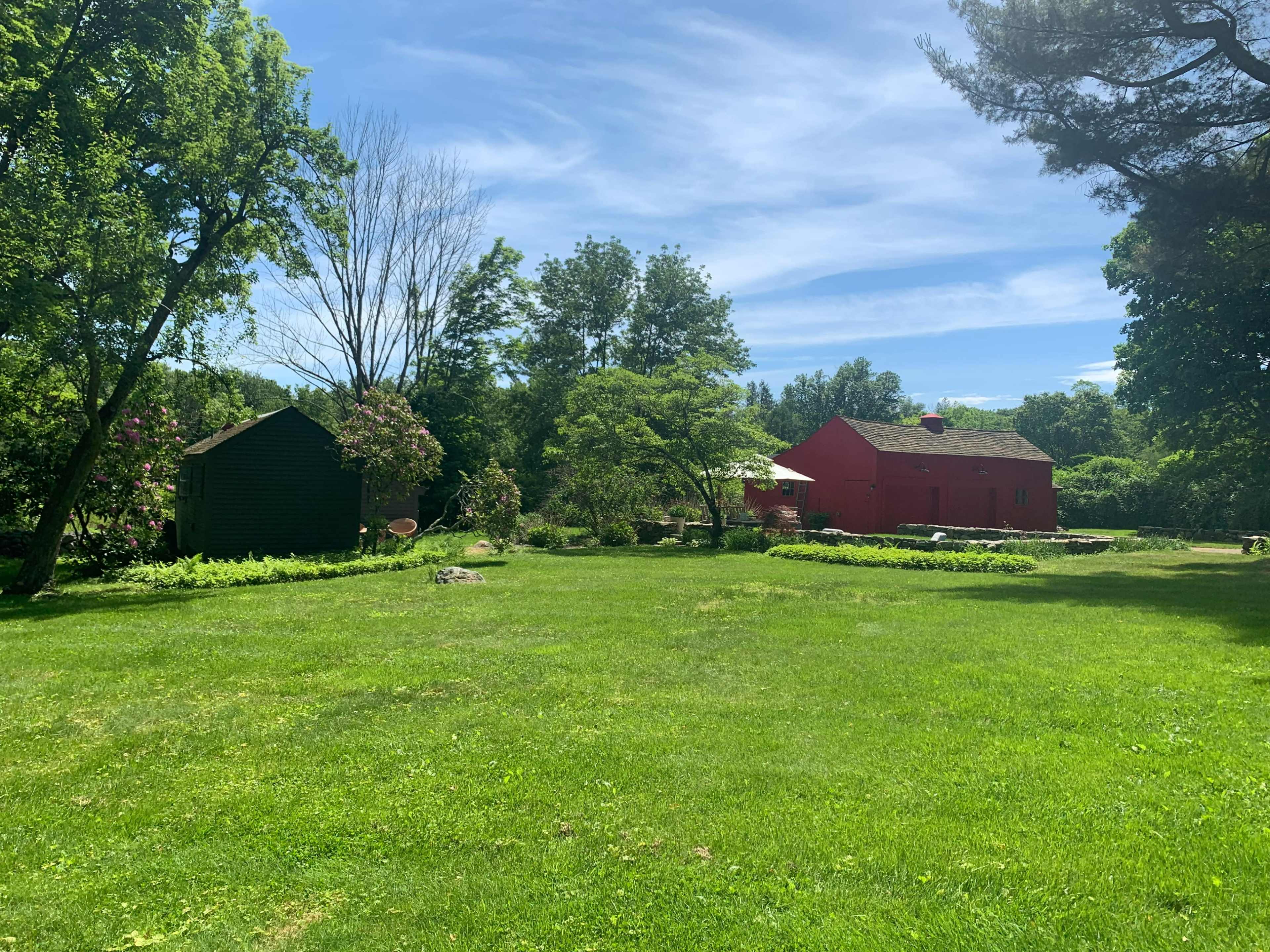 The image shows a green lawn with two buildings, one red and one dark, surrounded by trees and shrubs under a clear blue sky.