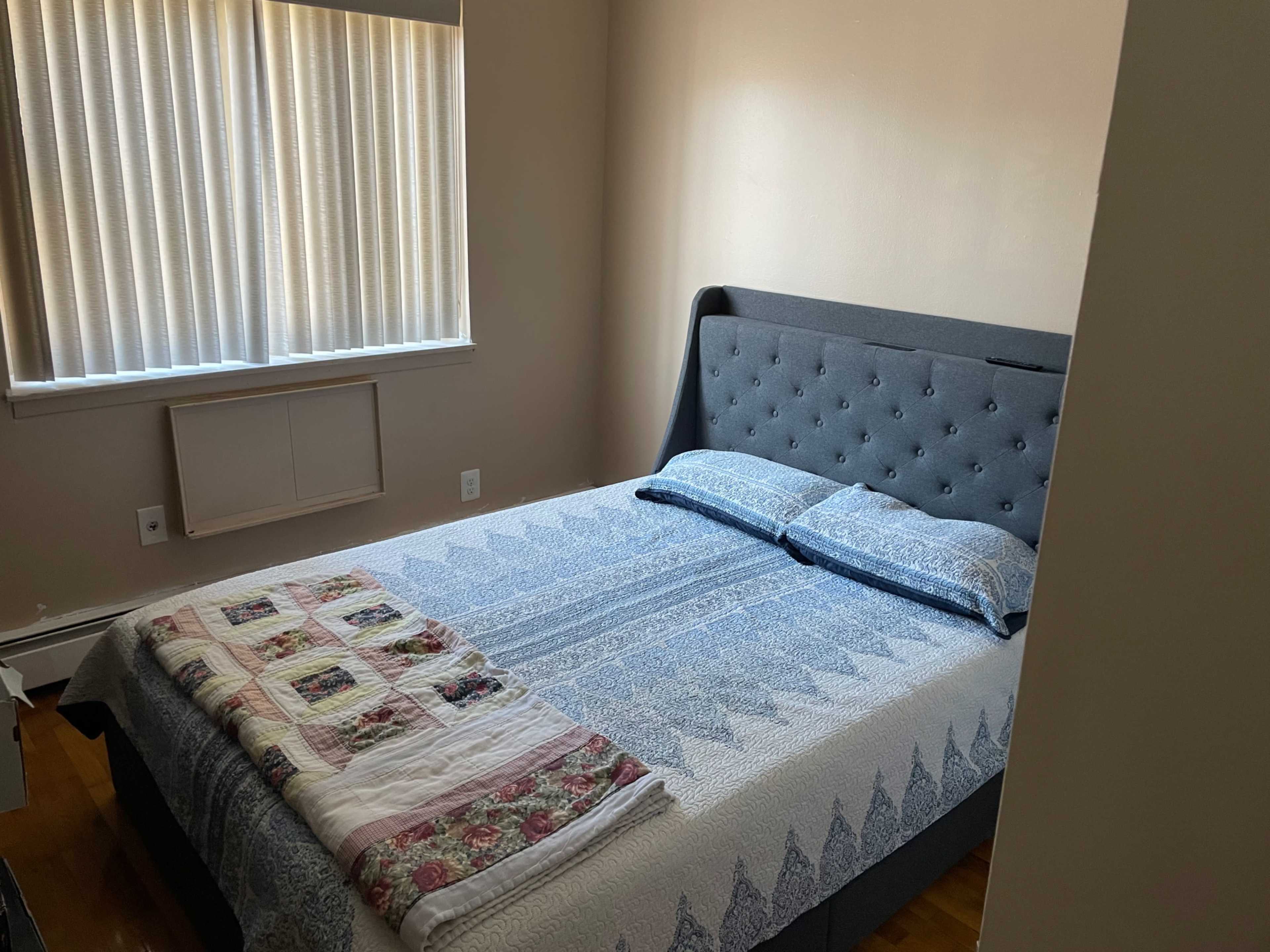 A small bedroom with a bed covered by a quilt, and sunlight filtering through vertical blinds on a window.