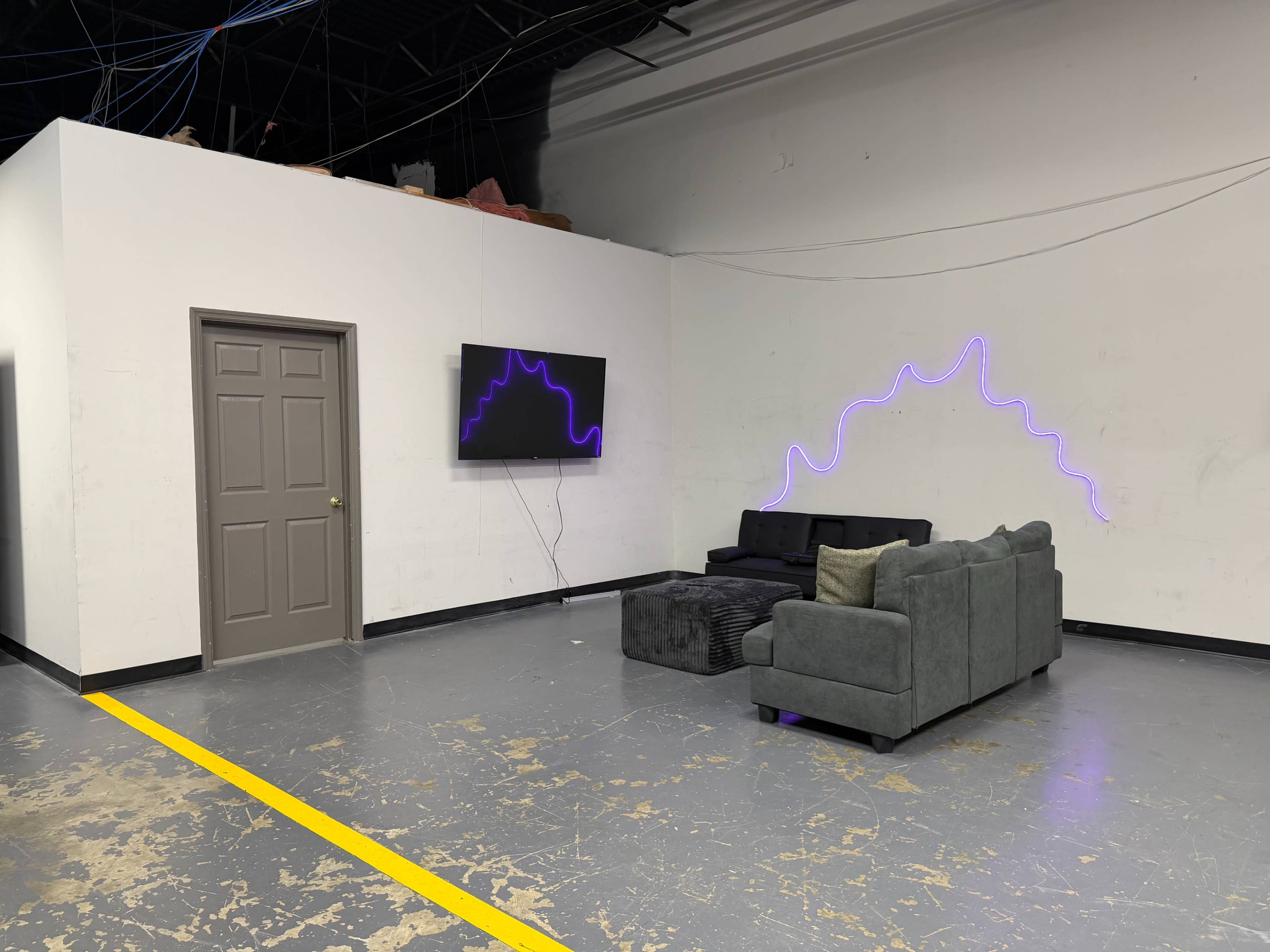 The image shows a minimalist interior space featuring a gray sectional sofa, a black ottoman, a wall-mounted television, and purple neon lighting curving along the wall.