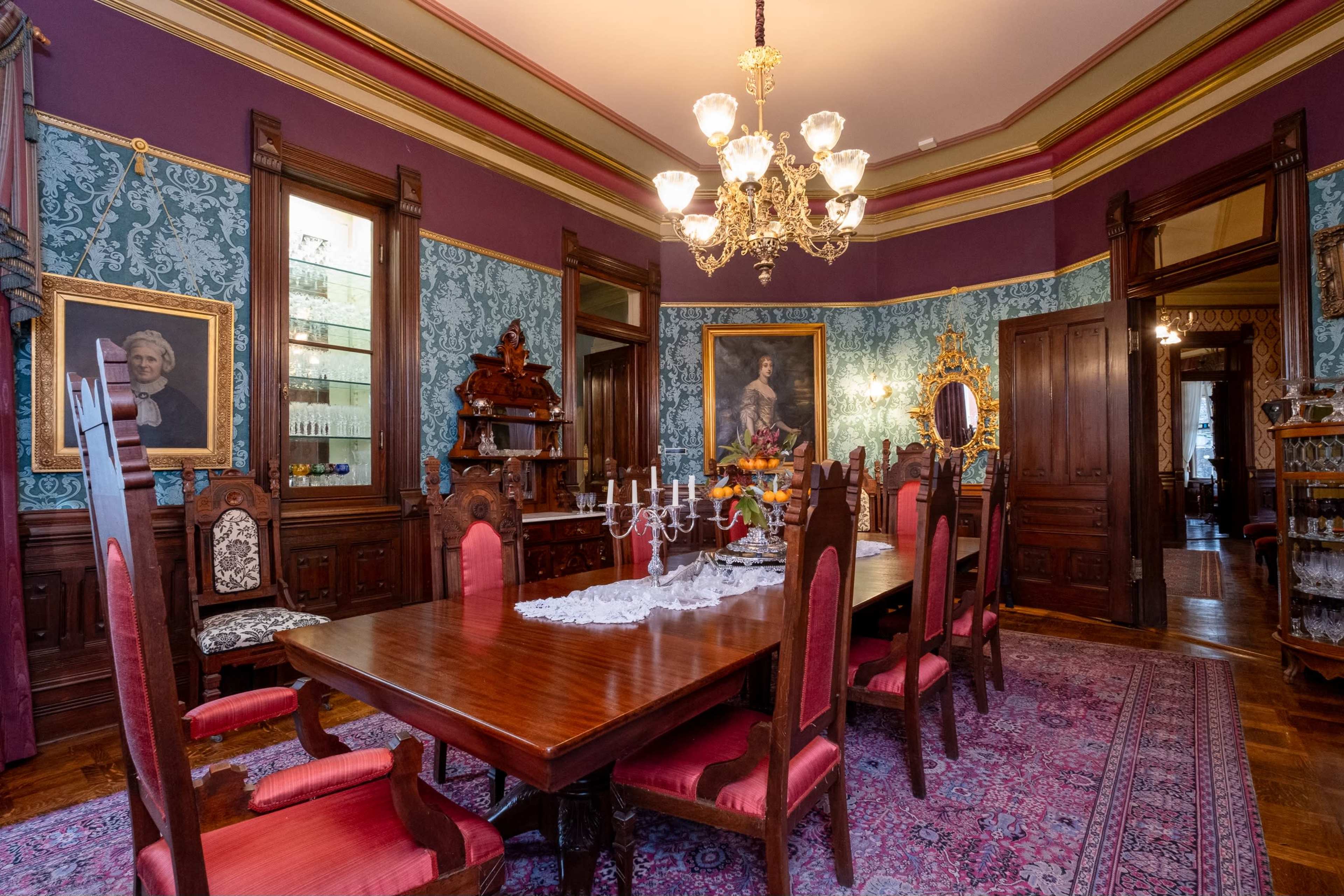 The image shows an ornate dining room featuring a long wooden table with red upholstered chairs, a chandelier, and decorative wallpaper.
