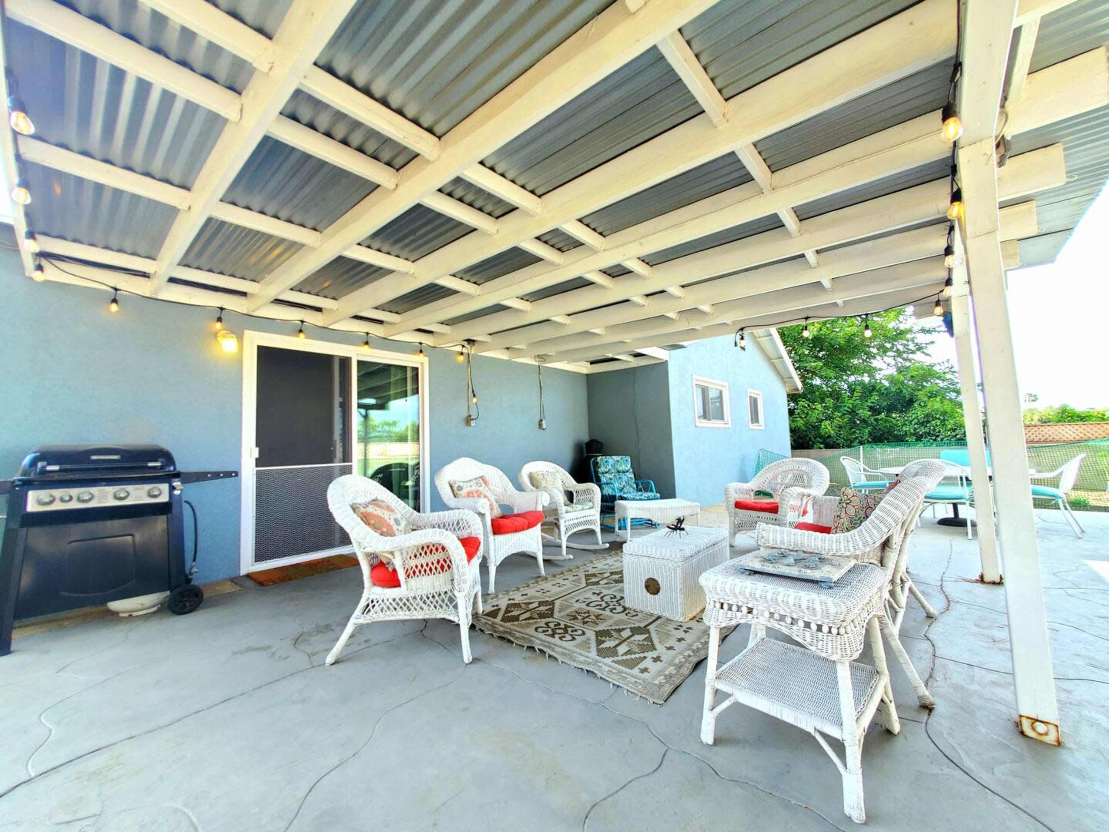 A covered patio area with white wicker chairs and a charcoal grill, surrounded by greenery.