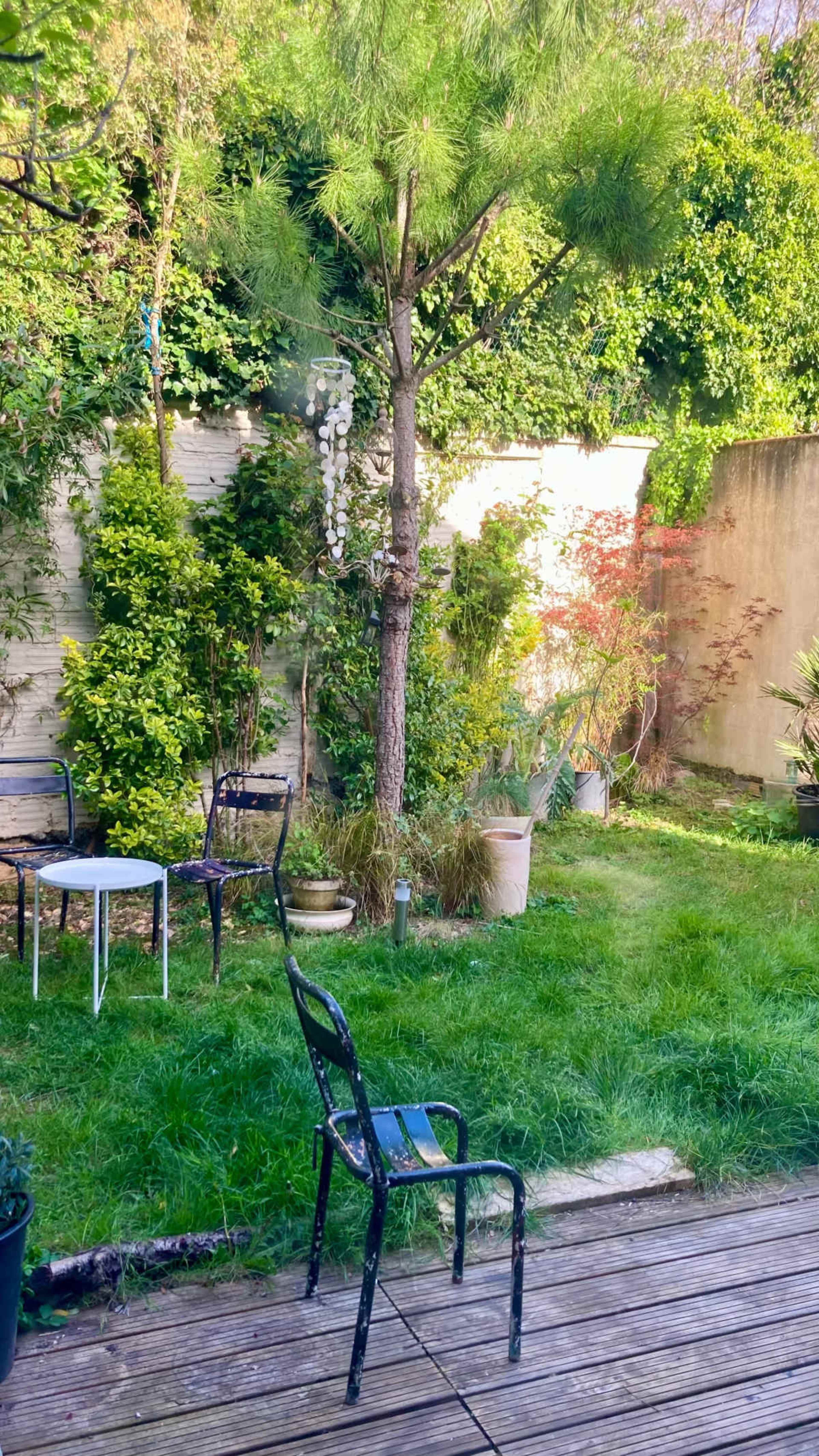 The image shows a garden with a green lawn, various plants and trees, and a wooden deck featuring a single black chair.