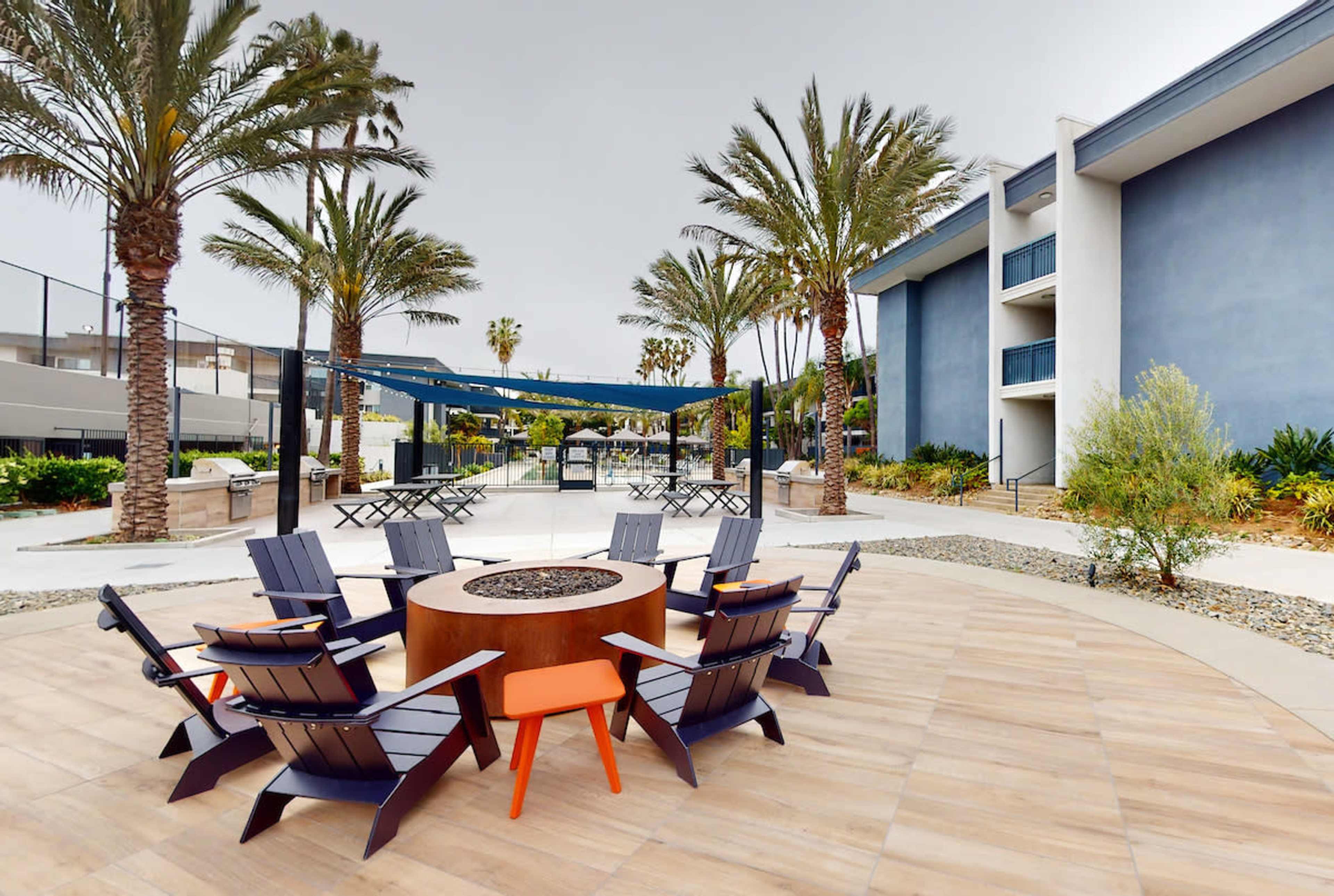 A landscaped outdoor area with a circular fire pit surrounded by eight wooden chairs, flanked by palm trees and a multi-story building in the background.