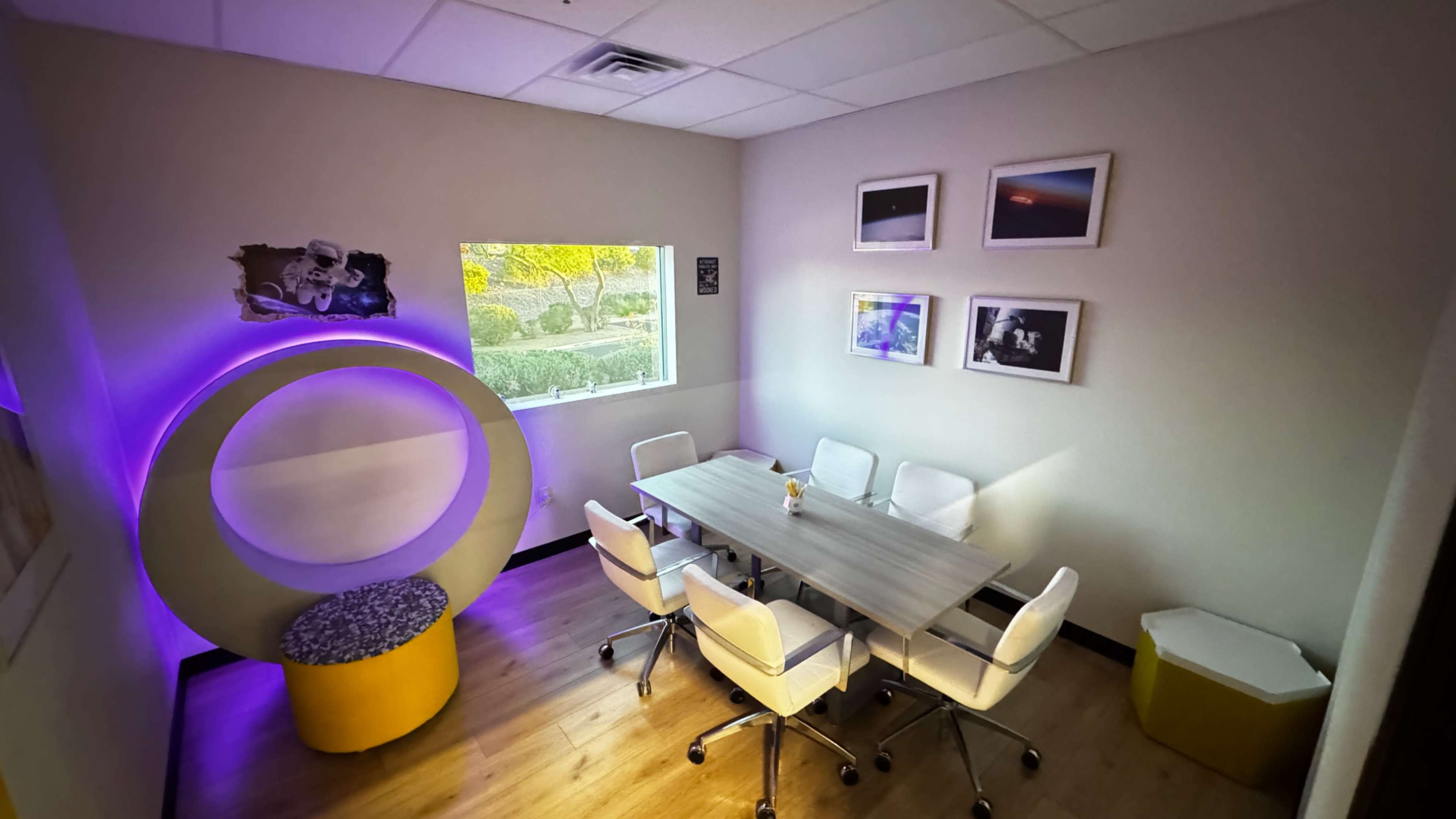 The image shows a modern conference room with a wooden table surrounded by white chairs, featuring a purple-lit circular seating area and framed photographs on the walls.