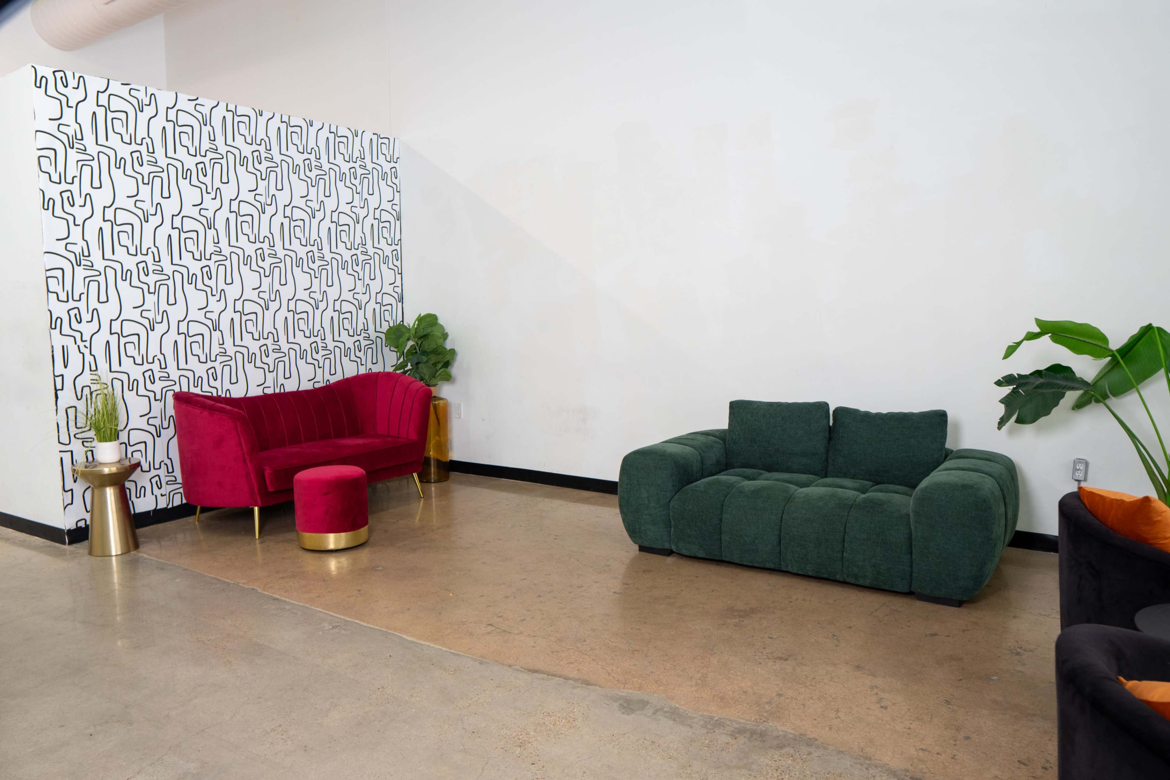 The image shows a modern interior space featuring a green couch, a pink velvet sofa, a small round pink table, and decorative plants against a patterned wall.