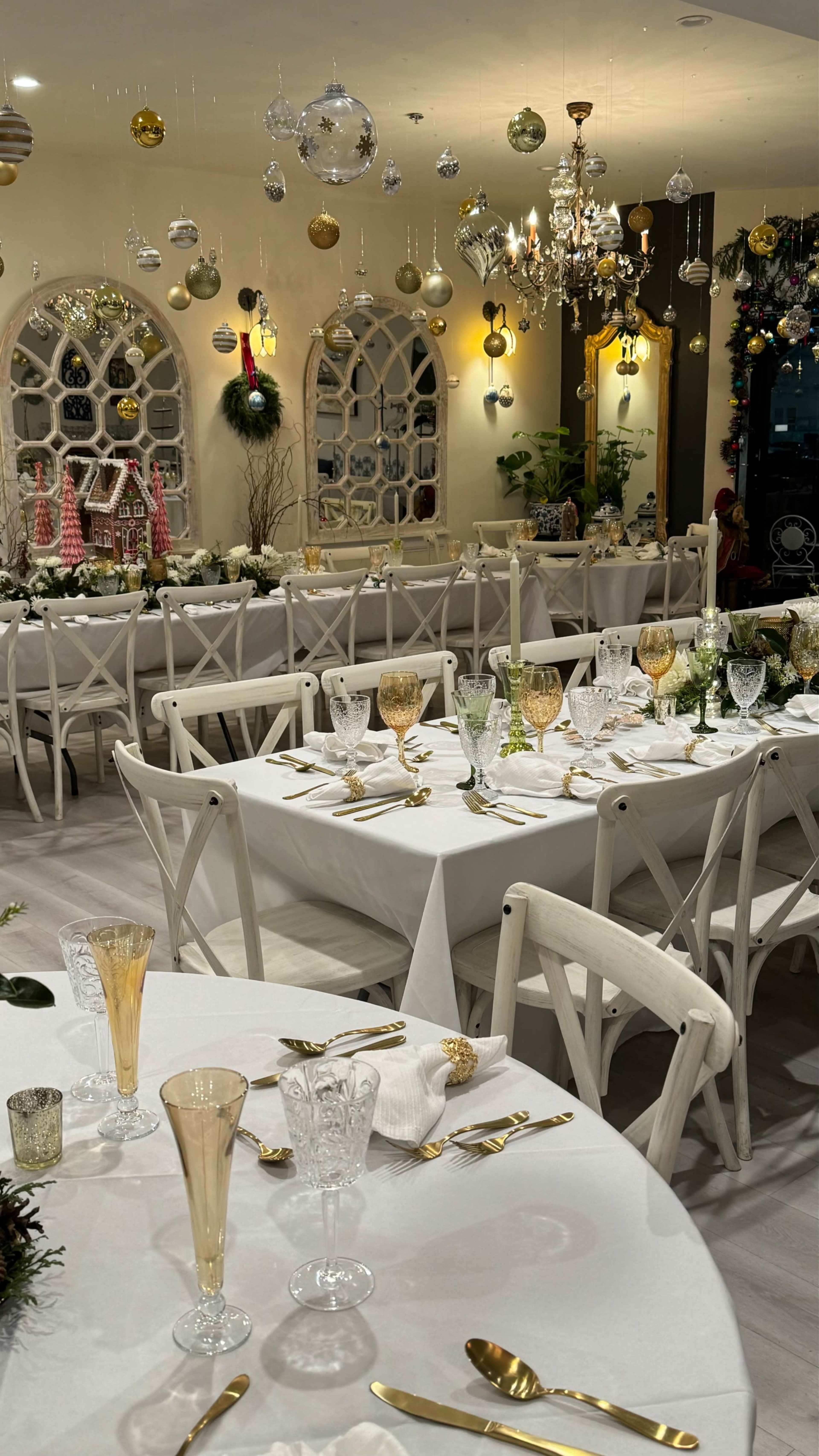 The image shows a decorated dining area with elegantly set tables, featuring white tablecloths, glassware, and hanging ornaments.