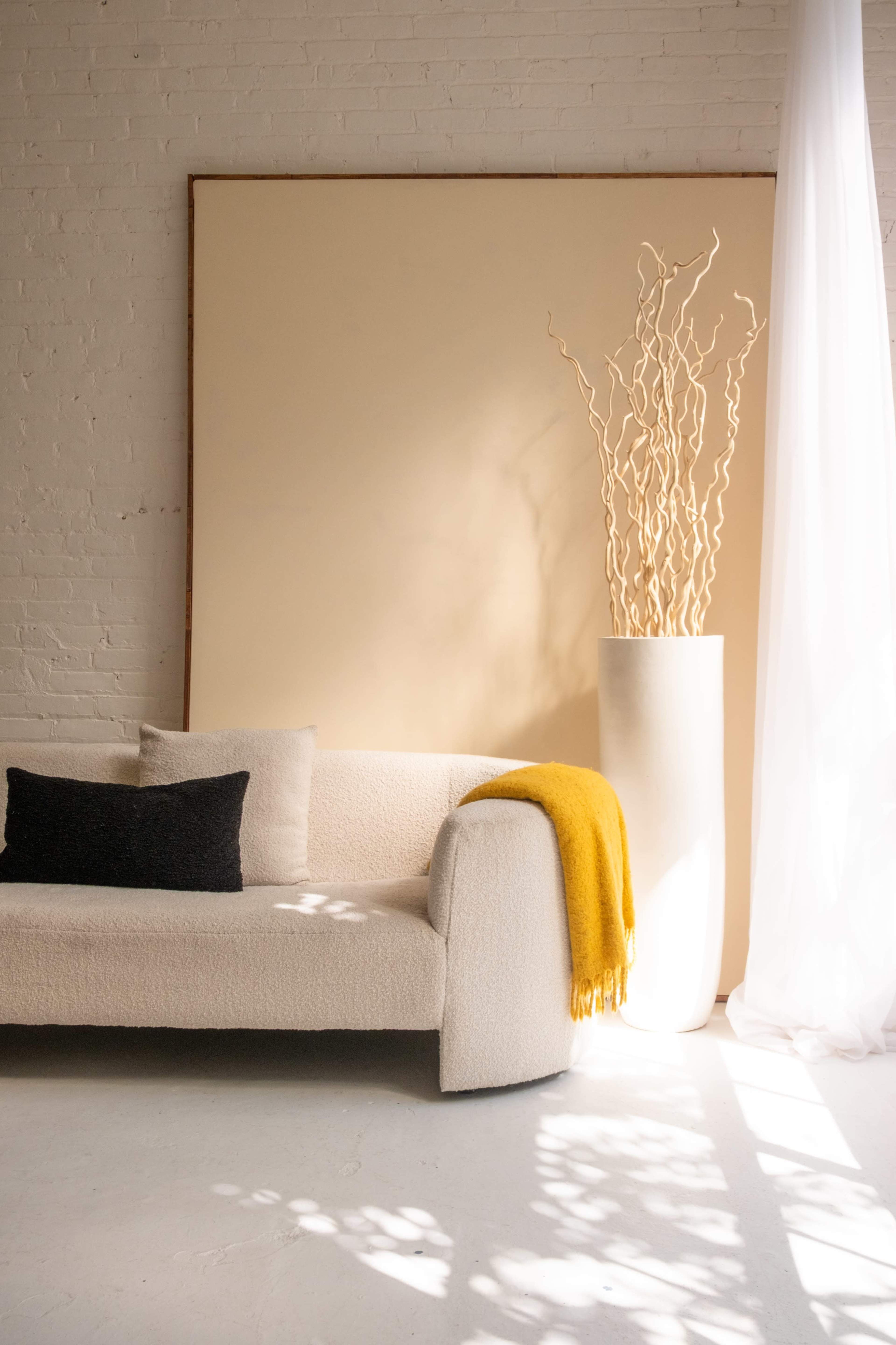 A light-filled interior features a textured sofa, a cylindrical vase with decorative branches, and a yellow throw draped over the arm.