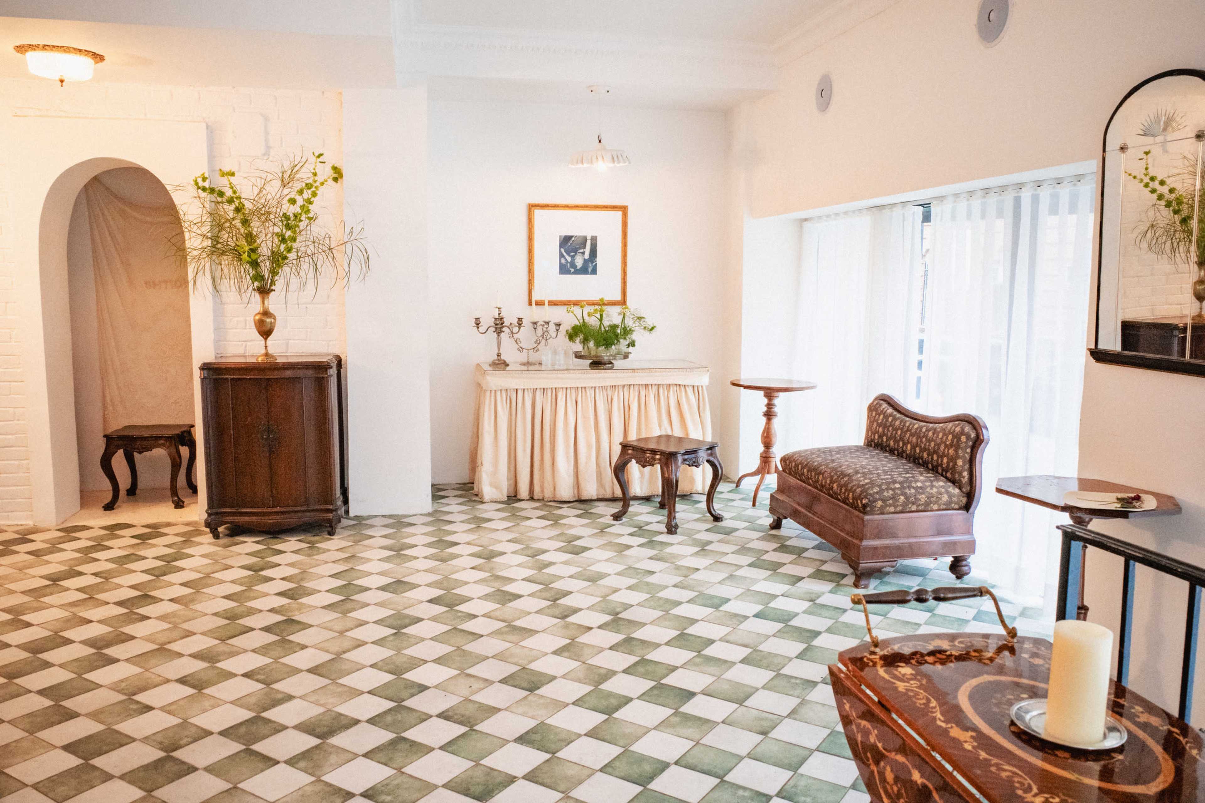 The image shows a spacious, well-lit room featuring vintage furniture, a floral arrangement, and a checkered floor.