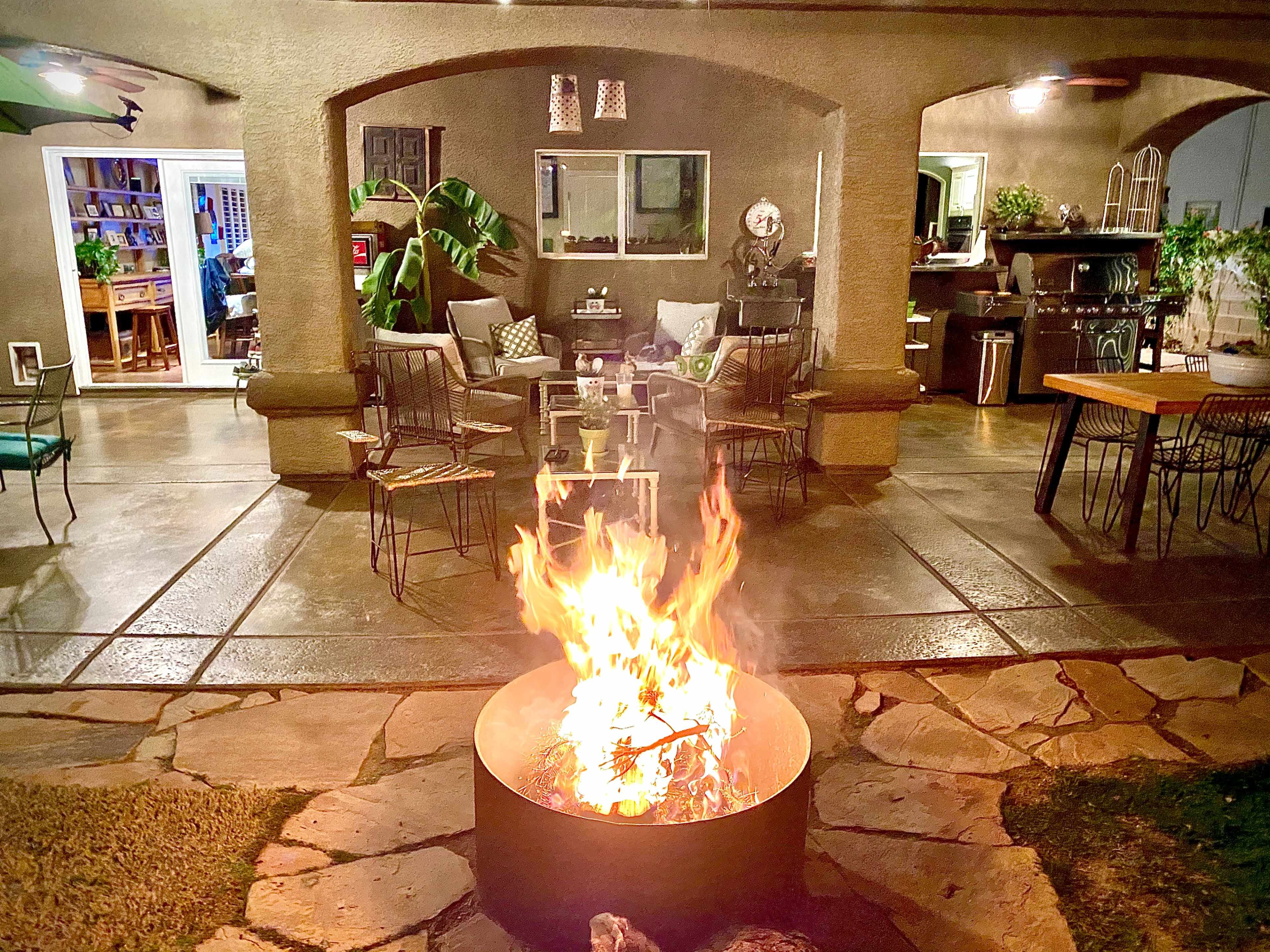 A fire pit burns brightly in the foreground of a patio area surrounded by chairs and tables.