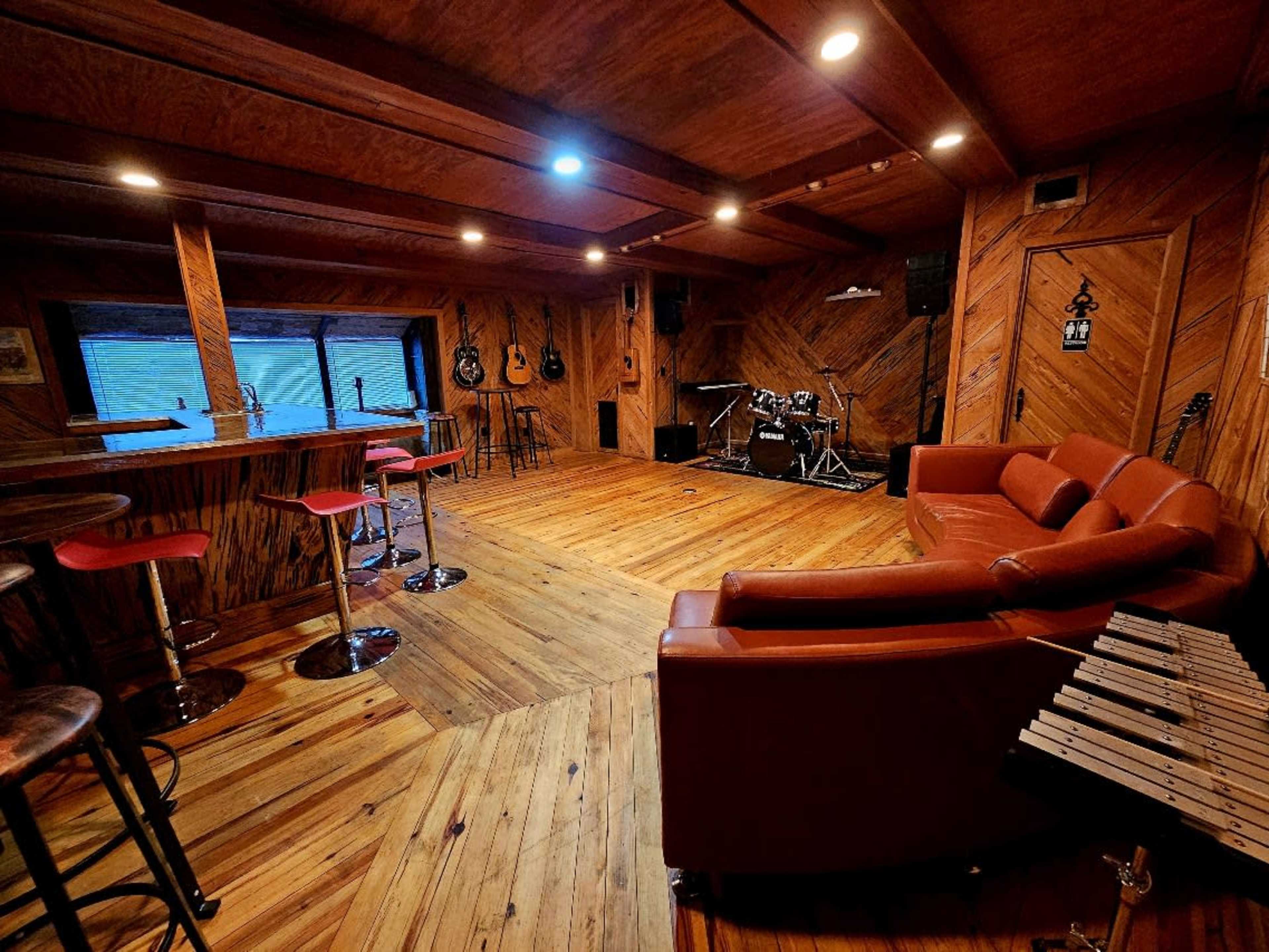 The image shows a cozy music rehearsal space with wooden walls, various musical instruments, and a seating area featuring a red couch and bar stools.
