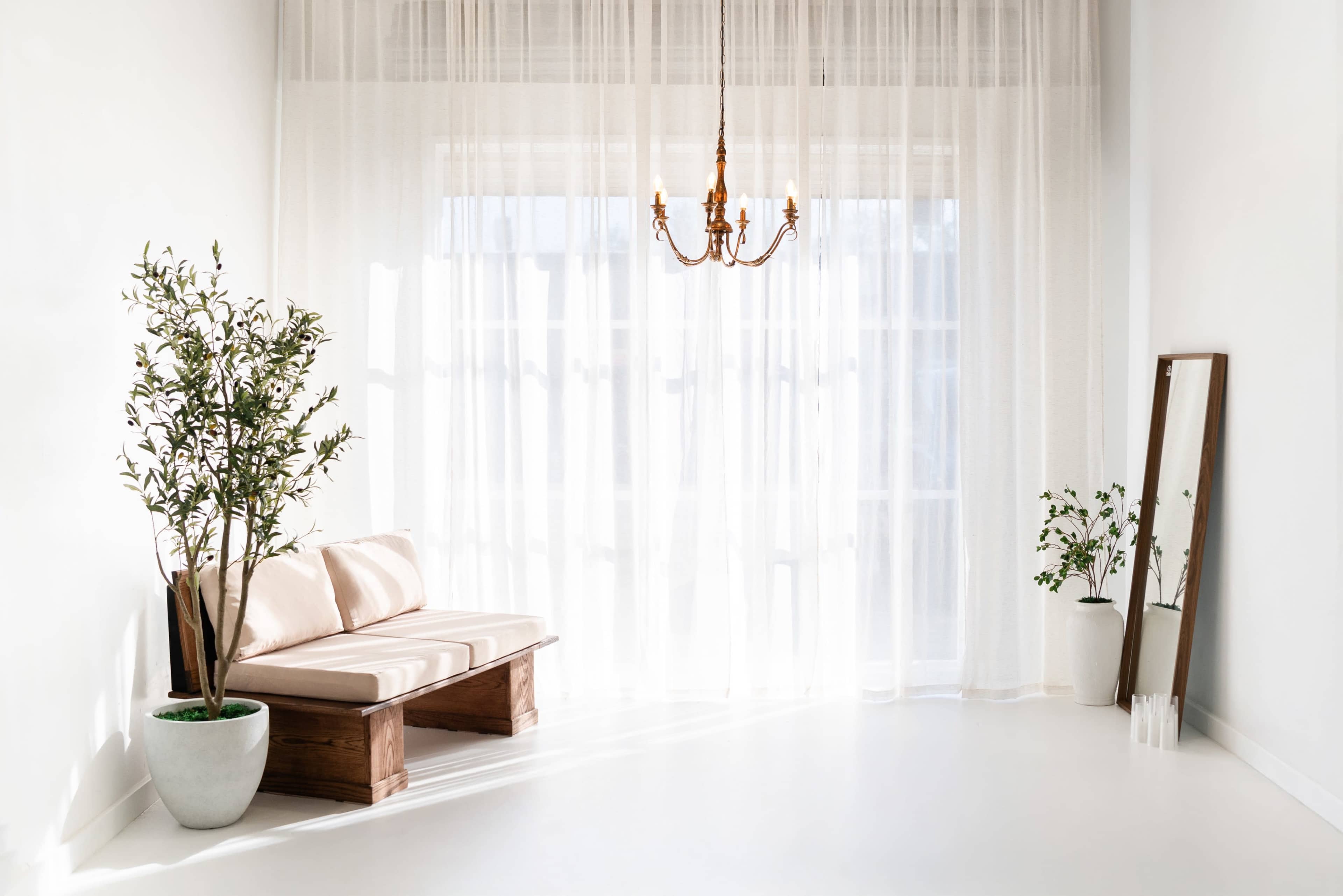 The image shows a minimalist indoor space with a wooden bench, a chandelier, potted plants, and sheer curtains allowing light to filter in.