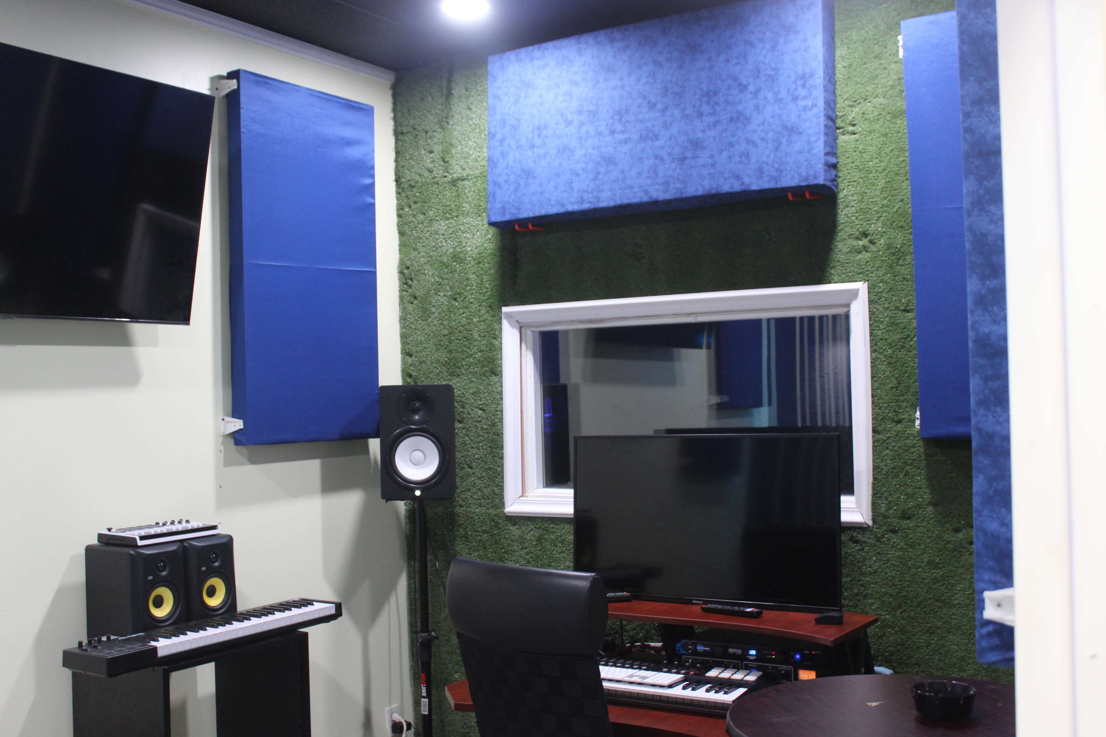 The image shows a recording studio with soundproofing panels on the walls, a monitor, speakers, and a keyboard setup.