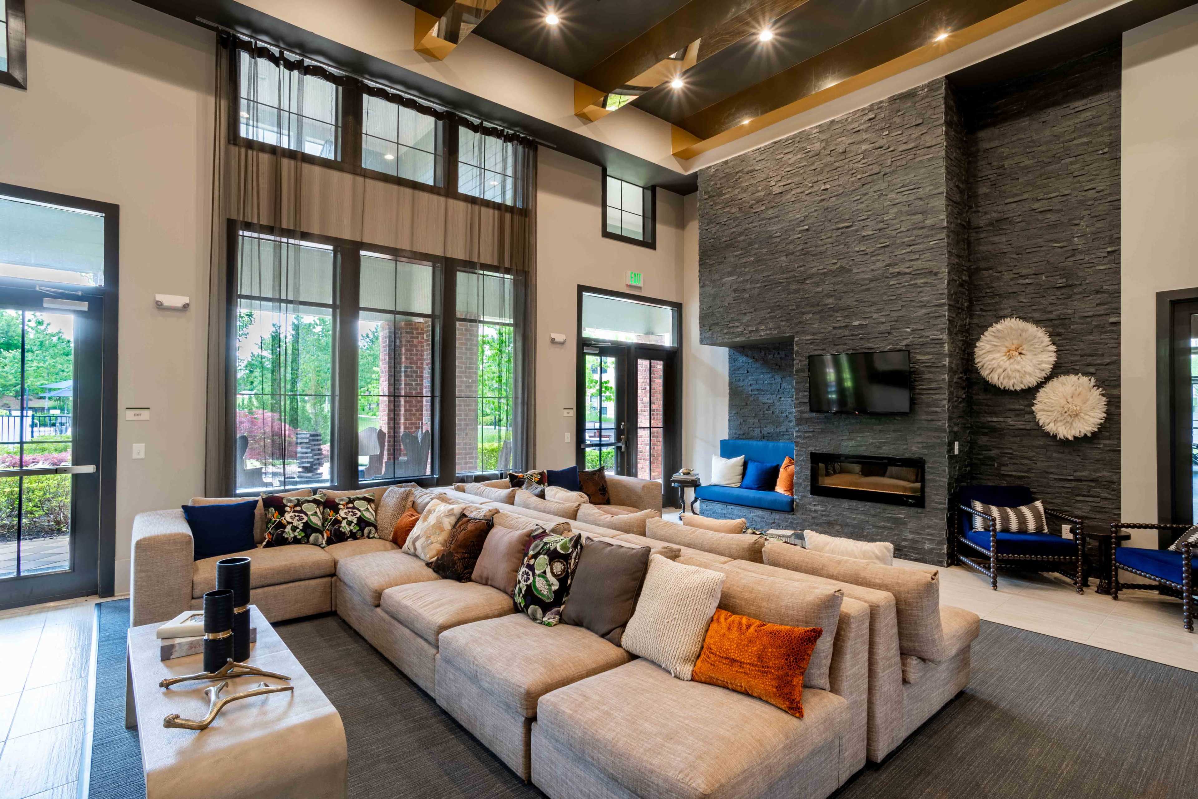 A modern lounge area featuring a large sectional sofa with decorative pillows, a stone fireplace, and large windows allowing natural light.