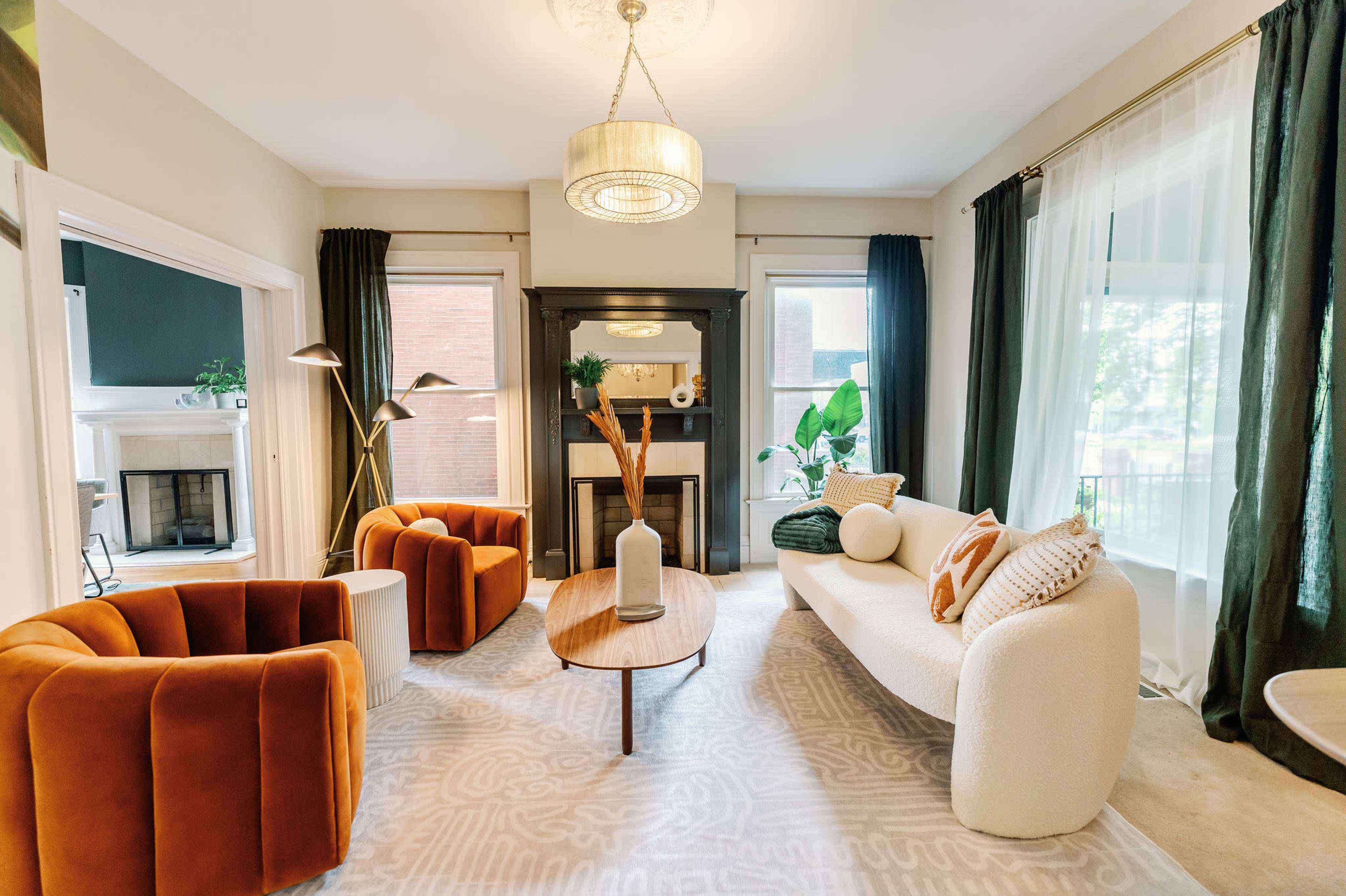A modern living room features two orange armchairs, a white sofa with decorative pillows, a round wooden coffee table, and large windows with green drapes.
