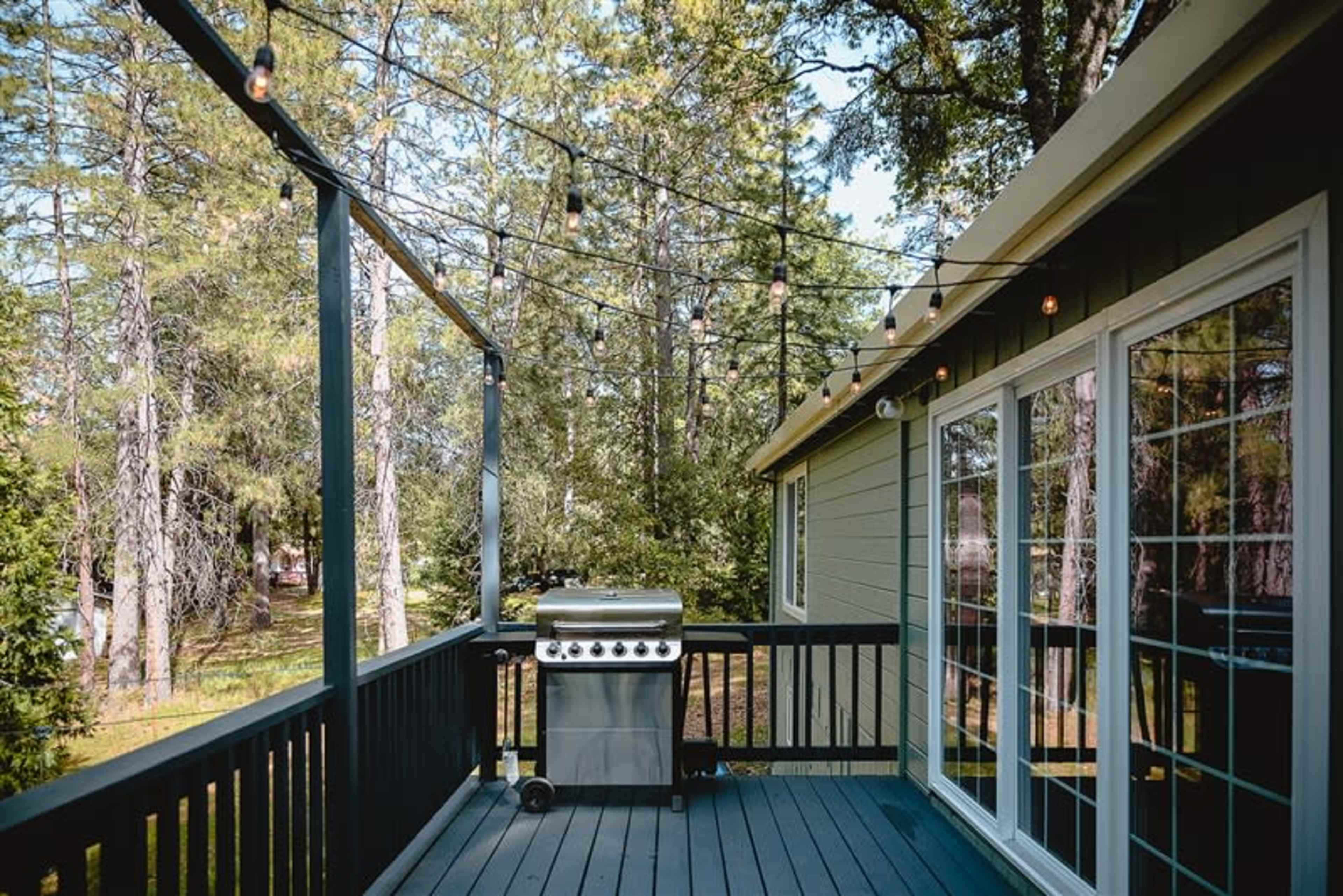 A wooden deck featuring a gas grill and string lights, surrounded by tall trees.