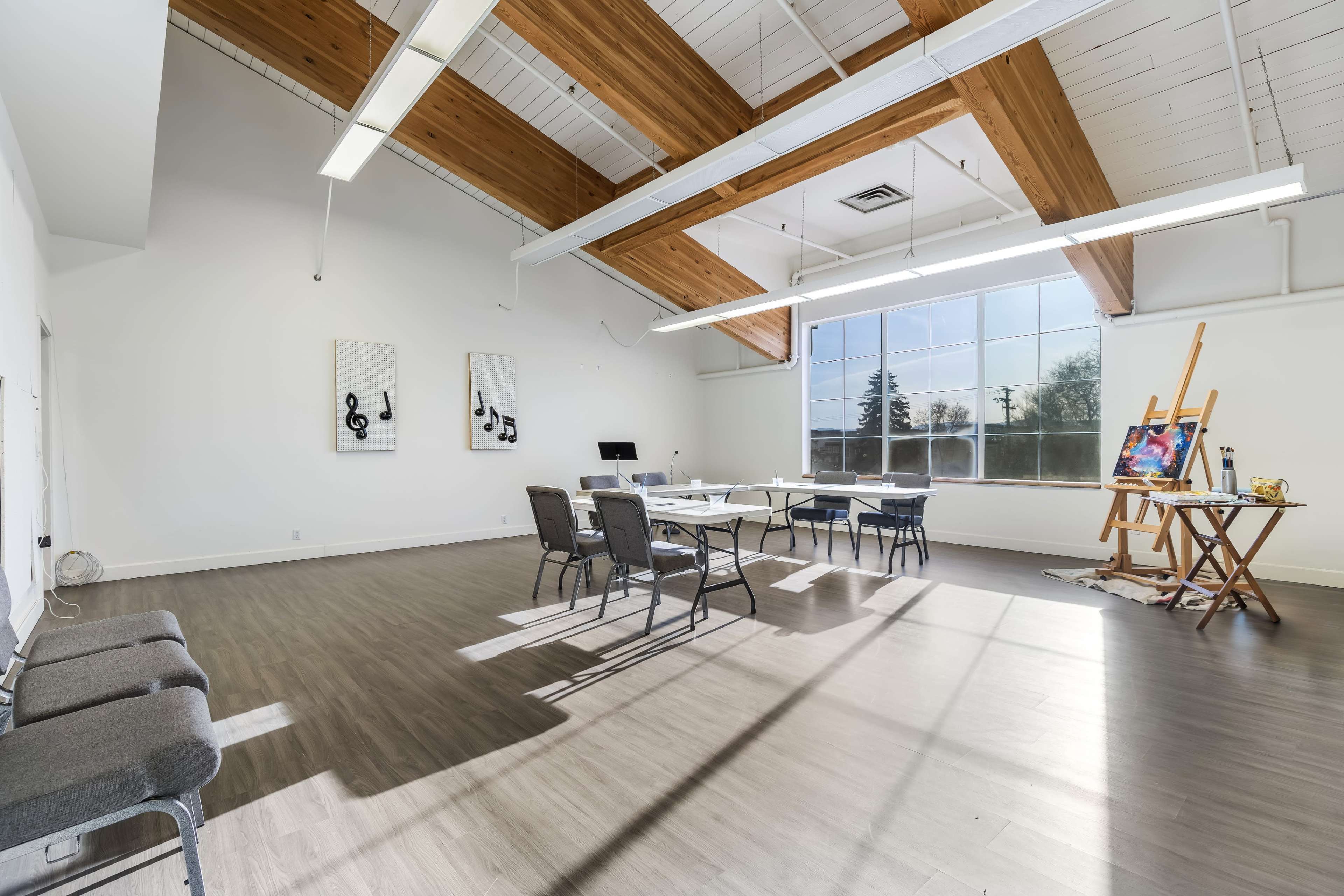 The image shows a spacious, well-lit room with high wooden beam ceilings, featuring tables and chairs arranged for a meeting or activity, along with an easel and some artwork displayed in the corner.