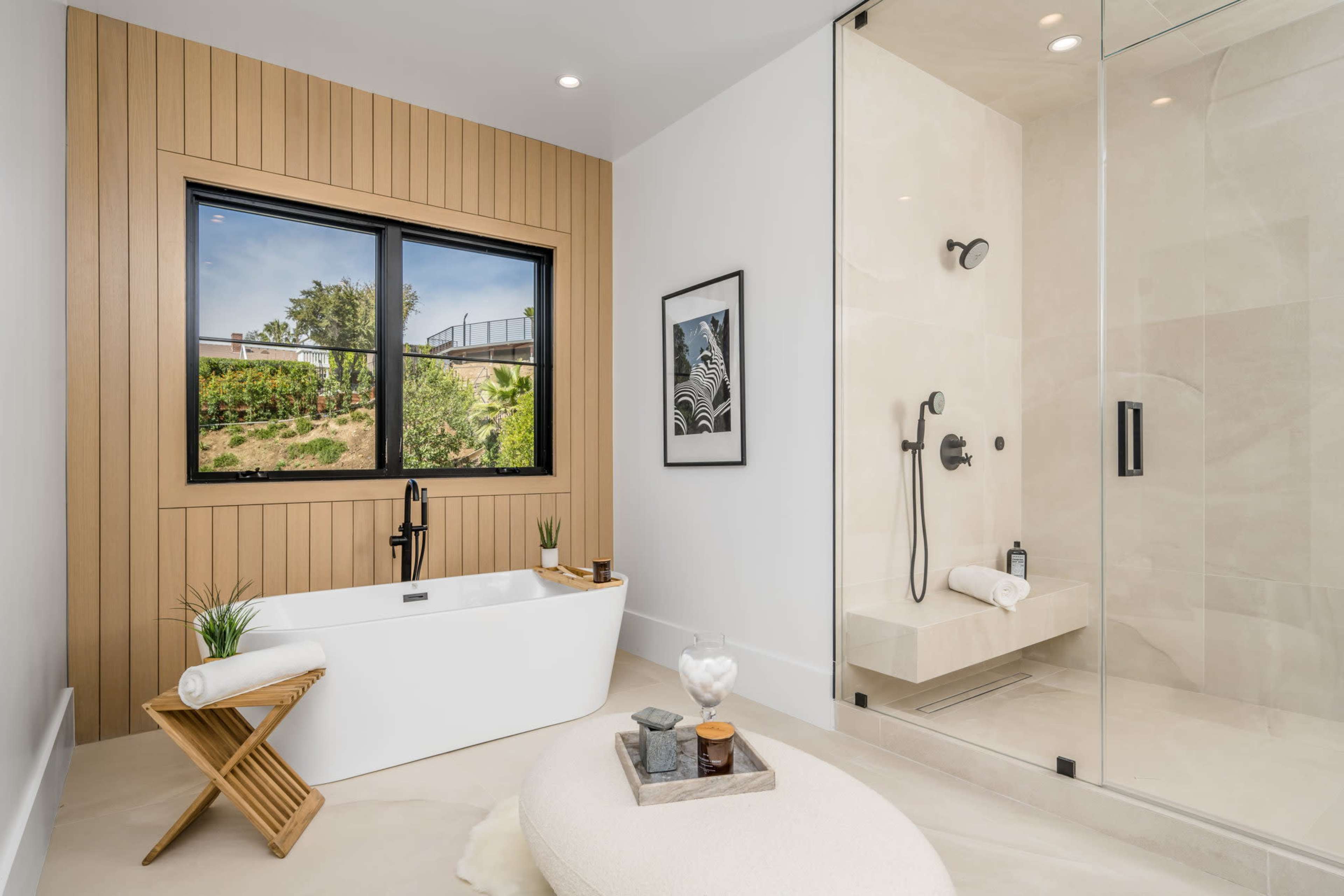 A modern bathroom featuring a freestanding bathtub, a glass shower enclosure, and large windows with a view of greenery.
