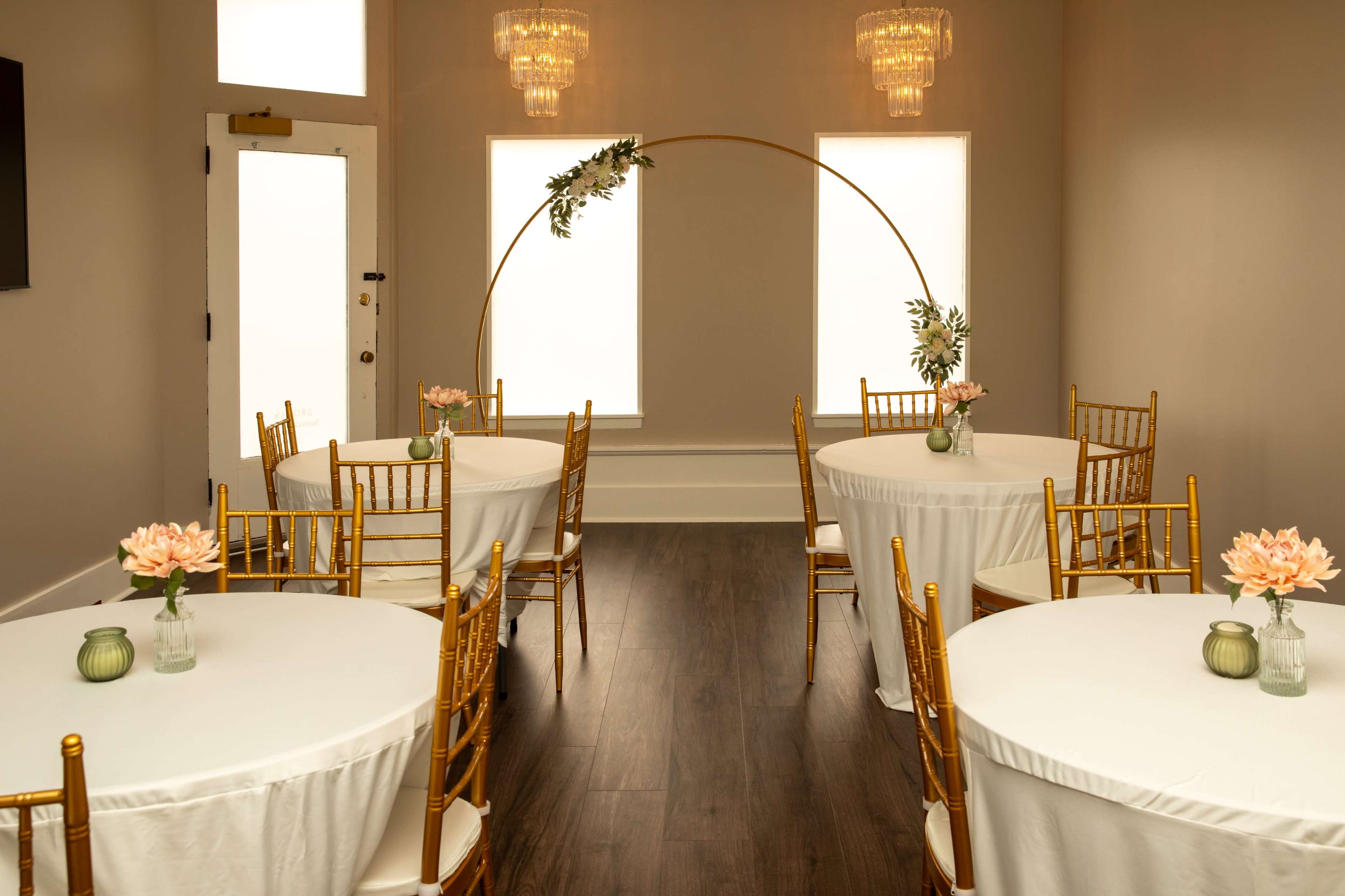 The image shows an elegantly arranged event space with round tables covered in white tablecloths, gold chairs, and a decorative arch with greenery and flowers.