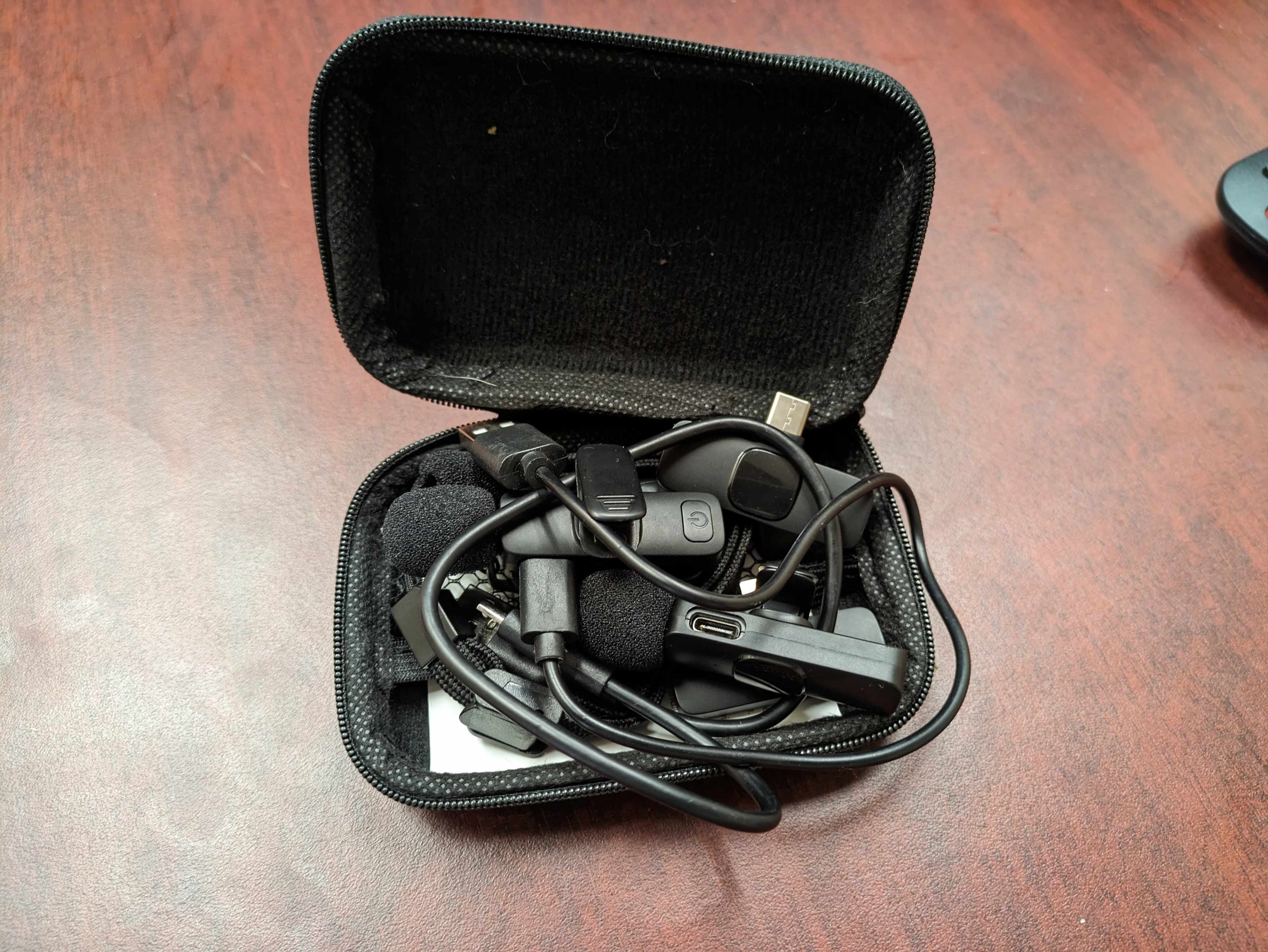 A black, zippered case contains tangled wires and various electronic accessories, including headphones and connectors, on a wooden surface.