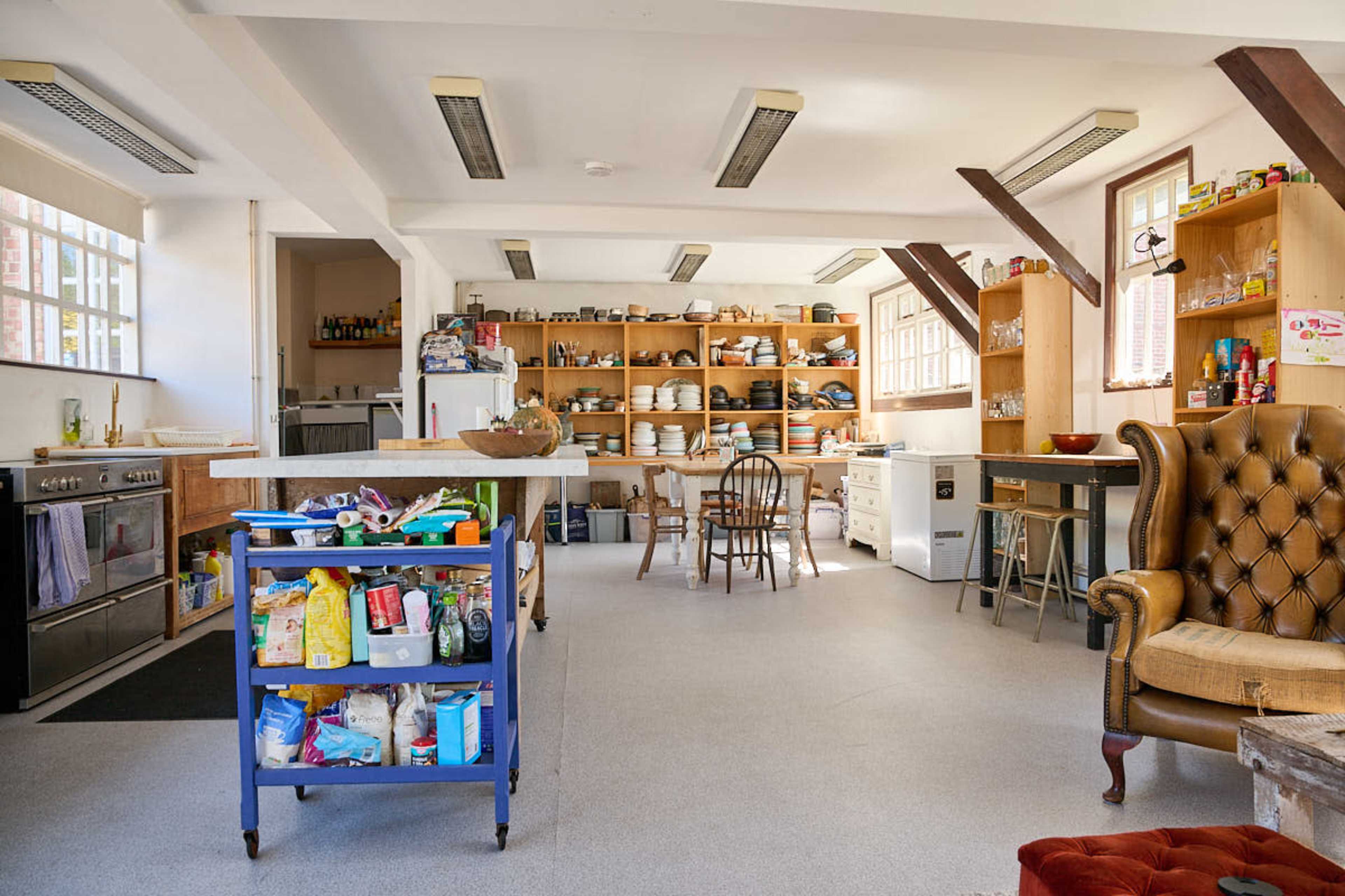 The image shows a spacious kitchen area with wooden shelves filled with dishes, a stove, a dining table, and a chair in a corner.