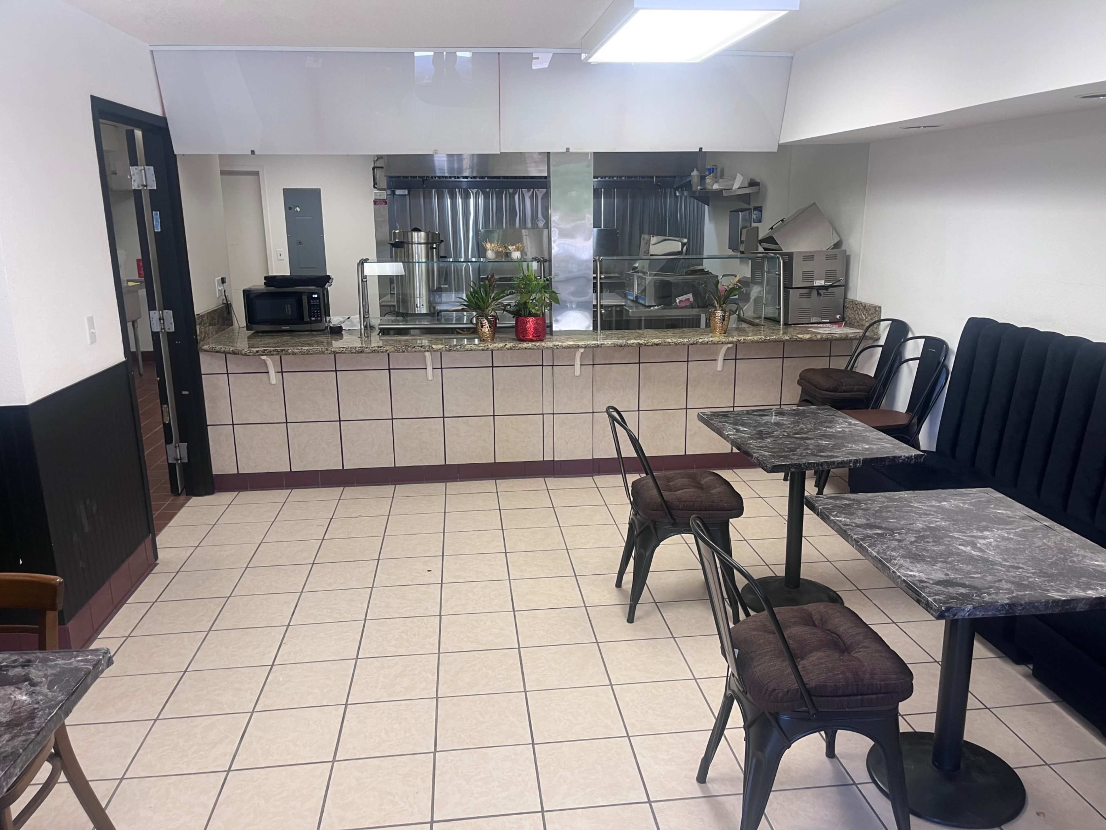The image shows a small, simple dining area with marble-topped tables and black metal chairs, leading to a kitchen behind a counter.