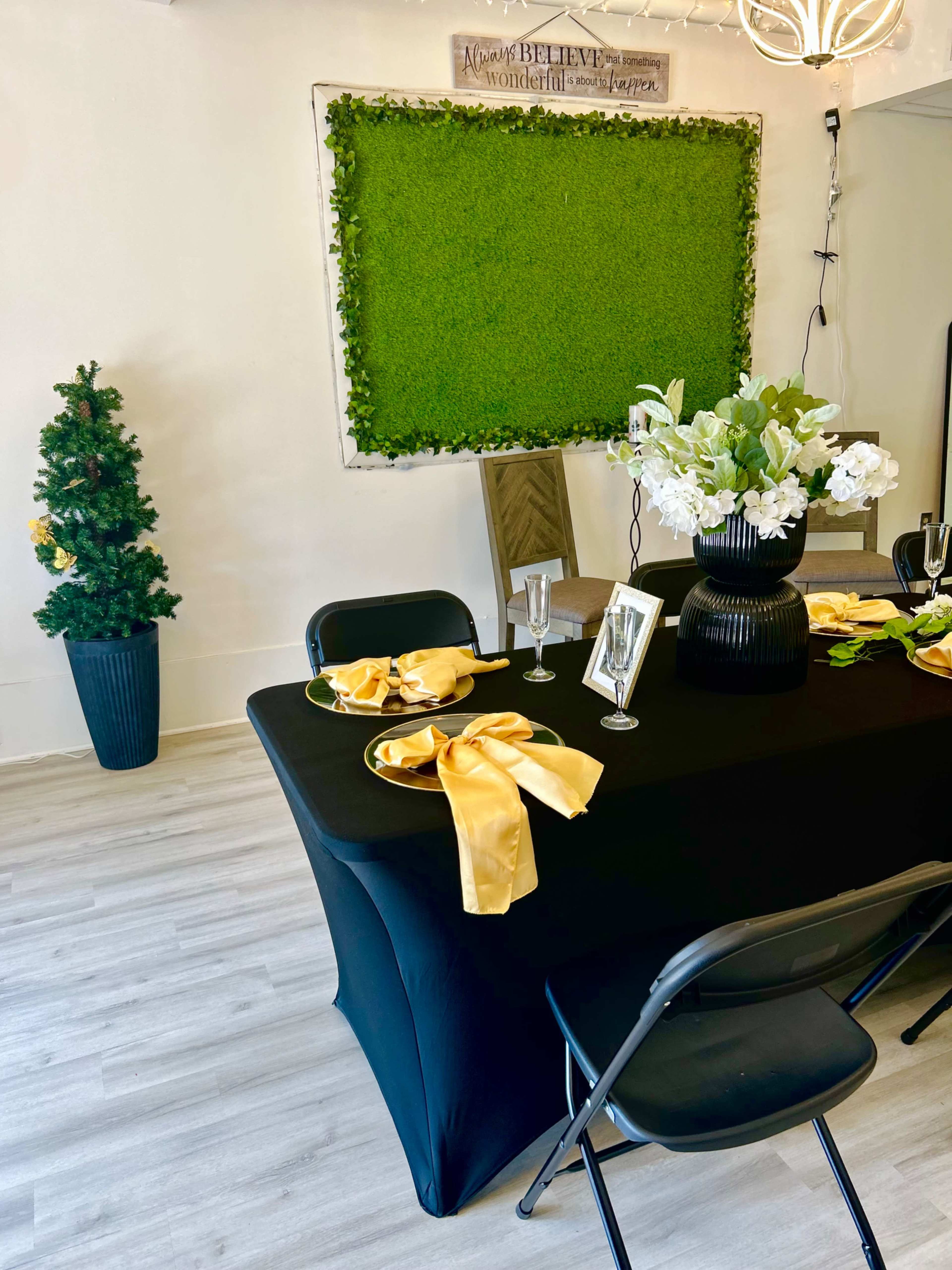 A black tablecloth covers a table set with yellow napkins and a vase of white flowers, positioned near a wall decorated with greenery and a small Christmas tree.