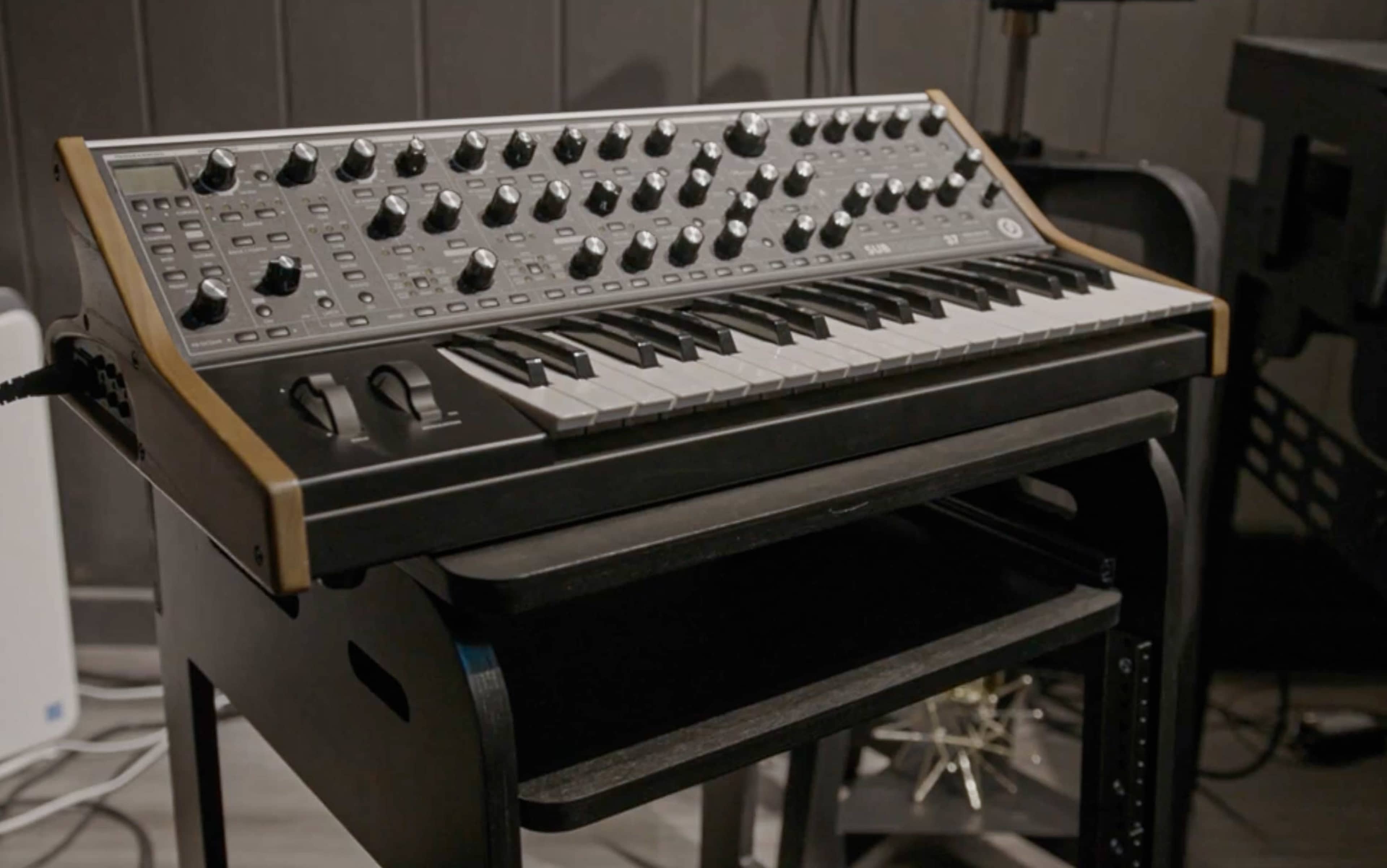 A synthesizer with a keyboard and control knobs is placed on a black stand in a music studio setting.