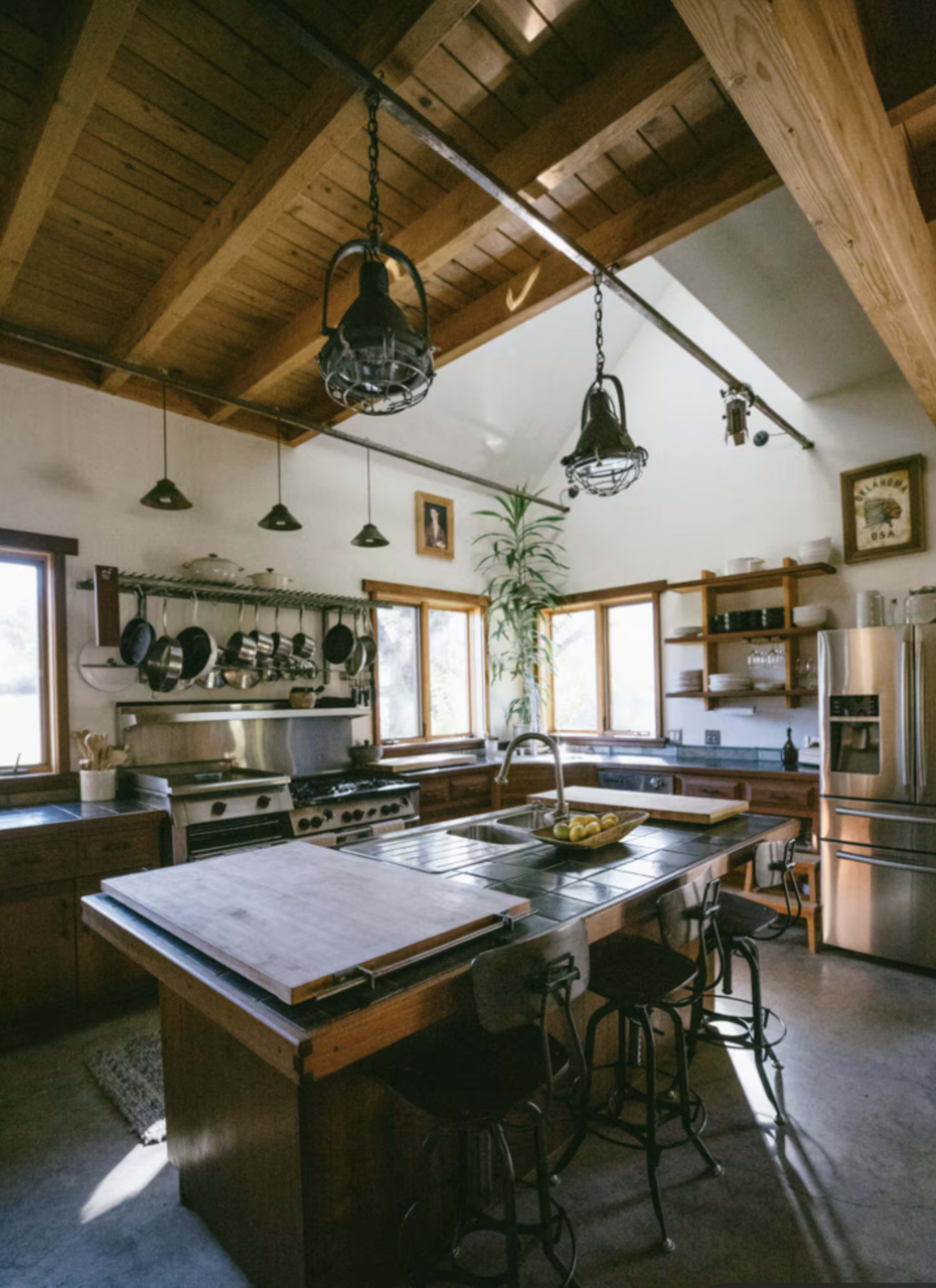 The image shows a rustic kitchen with wooden beams, an island with seating, and various cooking appliances and utensils arranged around it.