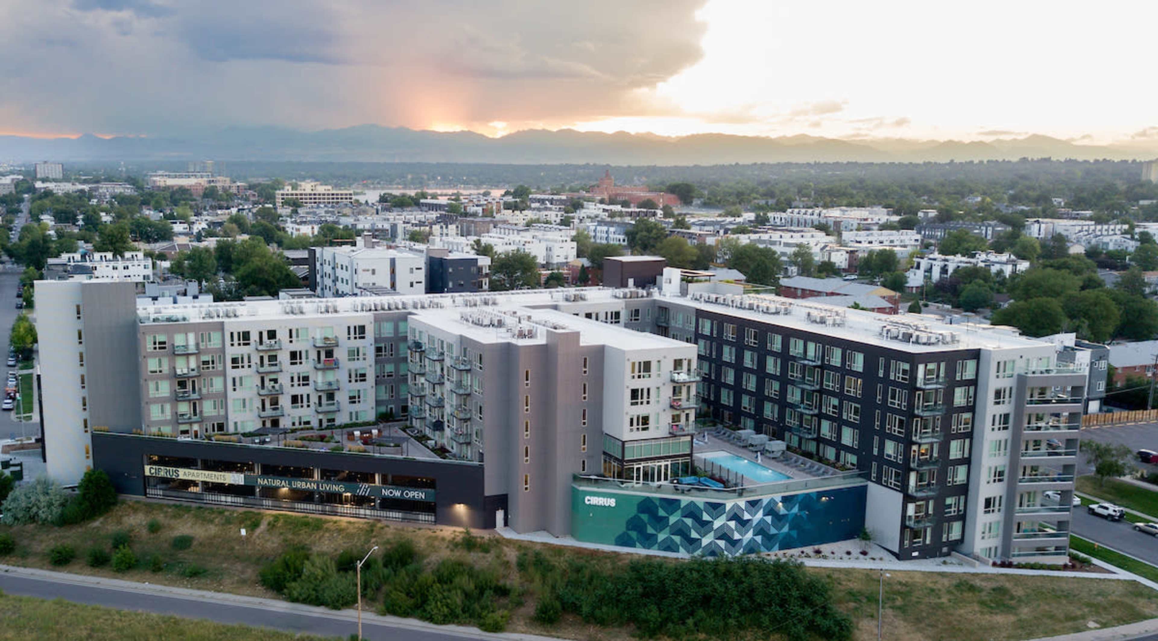 The image shows a modern apartment complex with a swimming pool, situated among a cityscape featuring residential buildings and distant mountains under a cloudy sky.