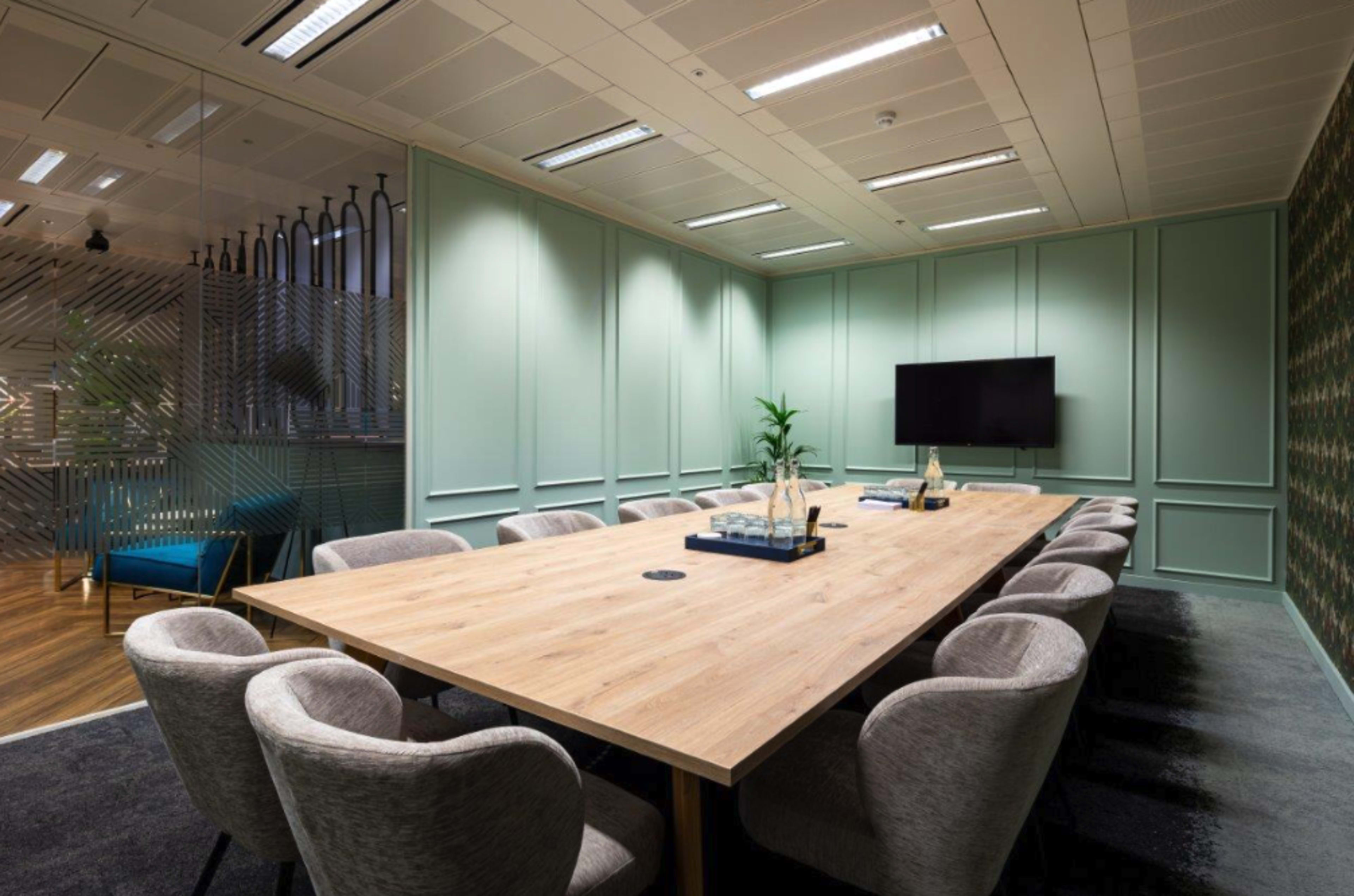 A modern conference room features a large wooden table surrounded by plush gray chairs, with green walls and a television on one side.