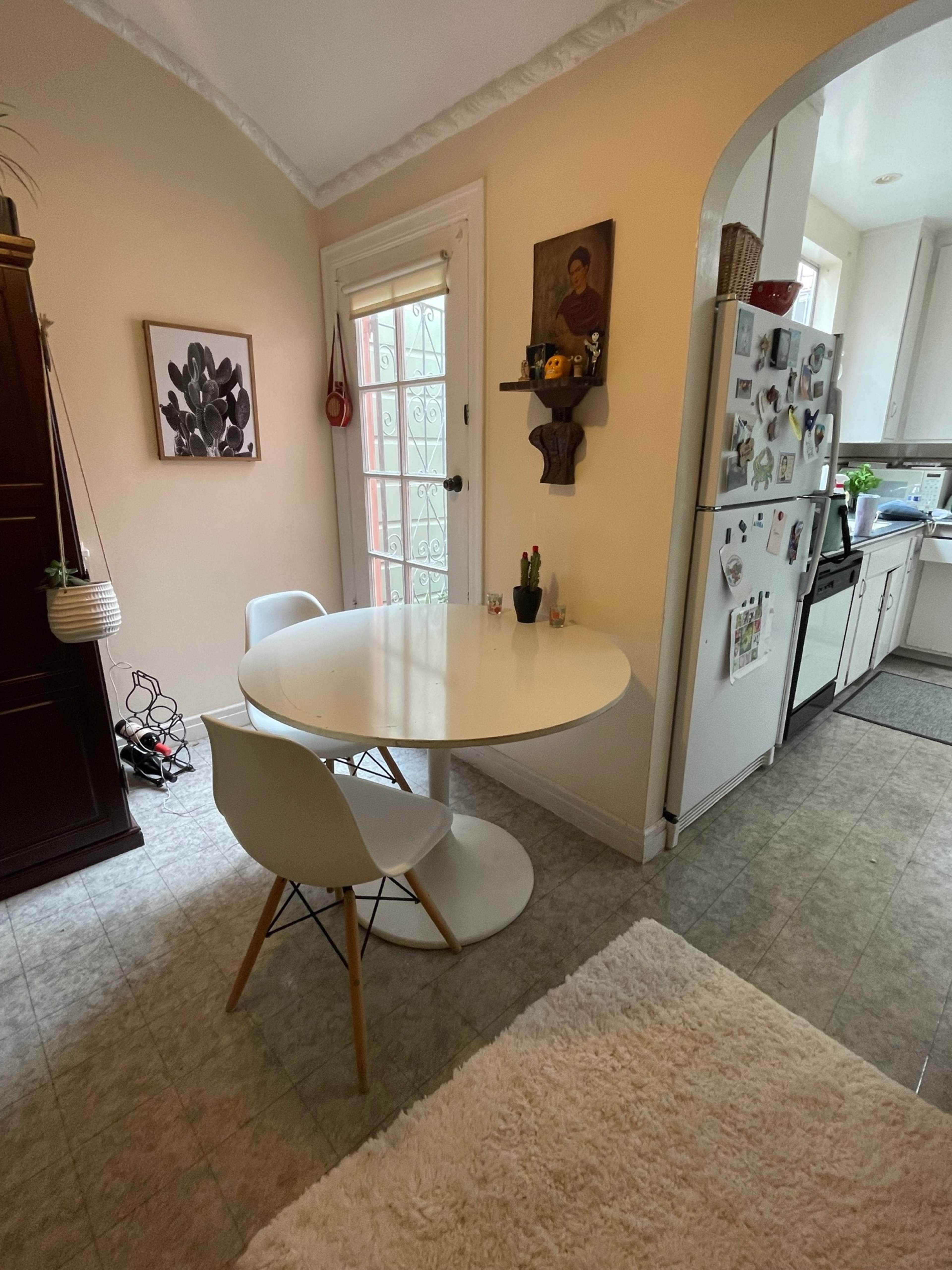 A small dining area features a round white table with two chairs beside a wall adorned with artwork, and a kitchen visible in the background.