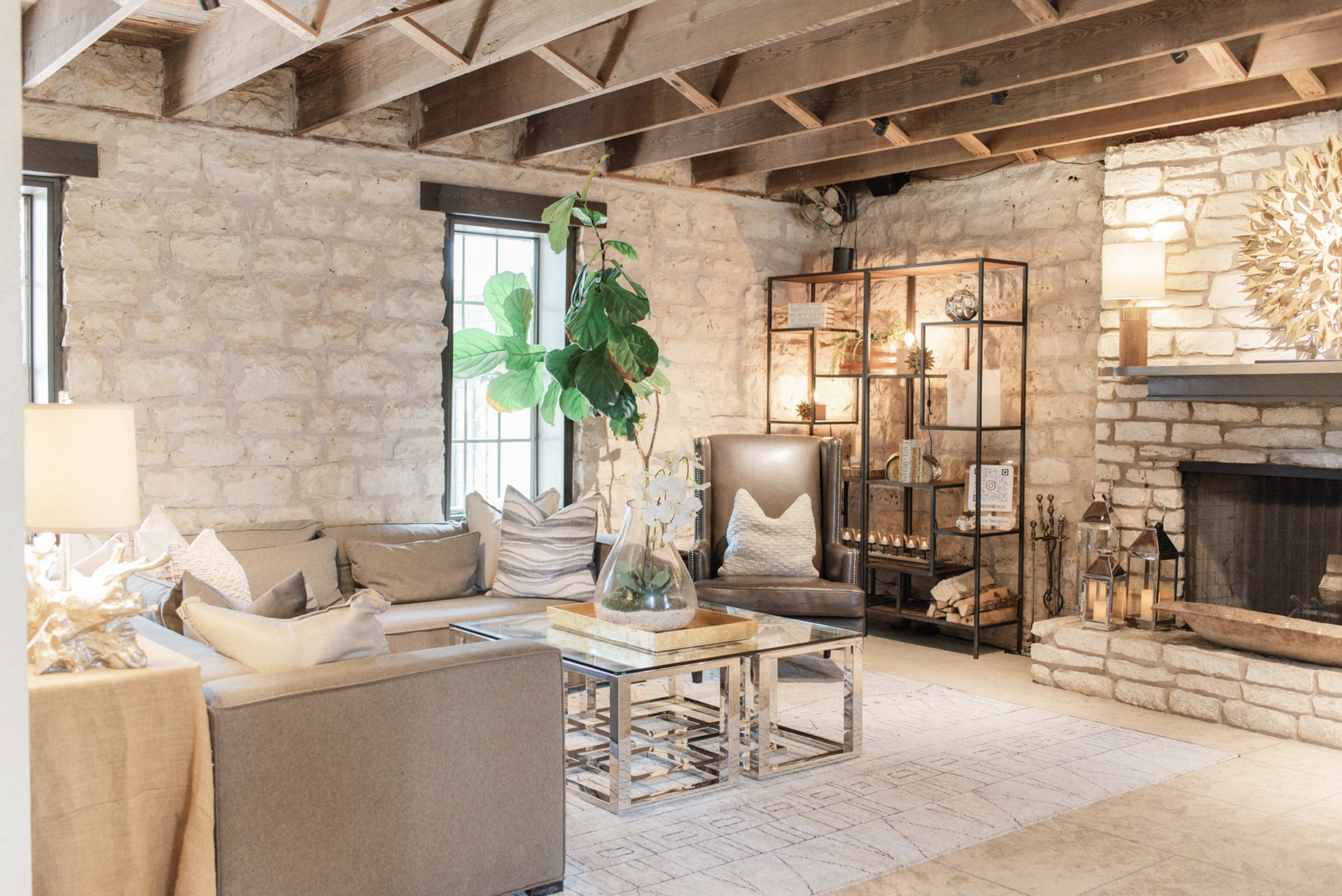 The image shows a cozy living room with stone walls, a fireplace, a sectional sofa, and a decorative bookshelf.
