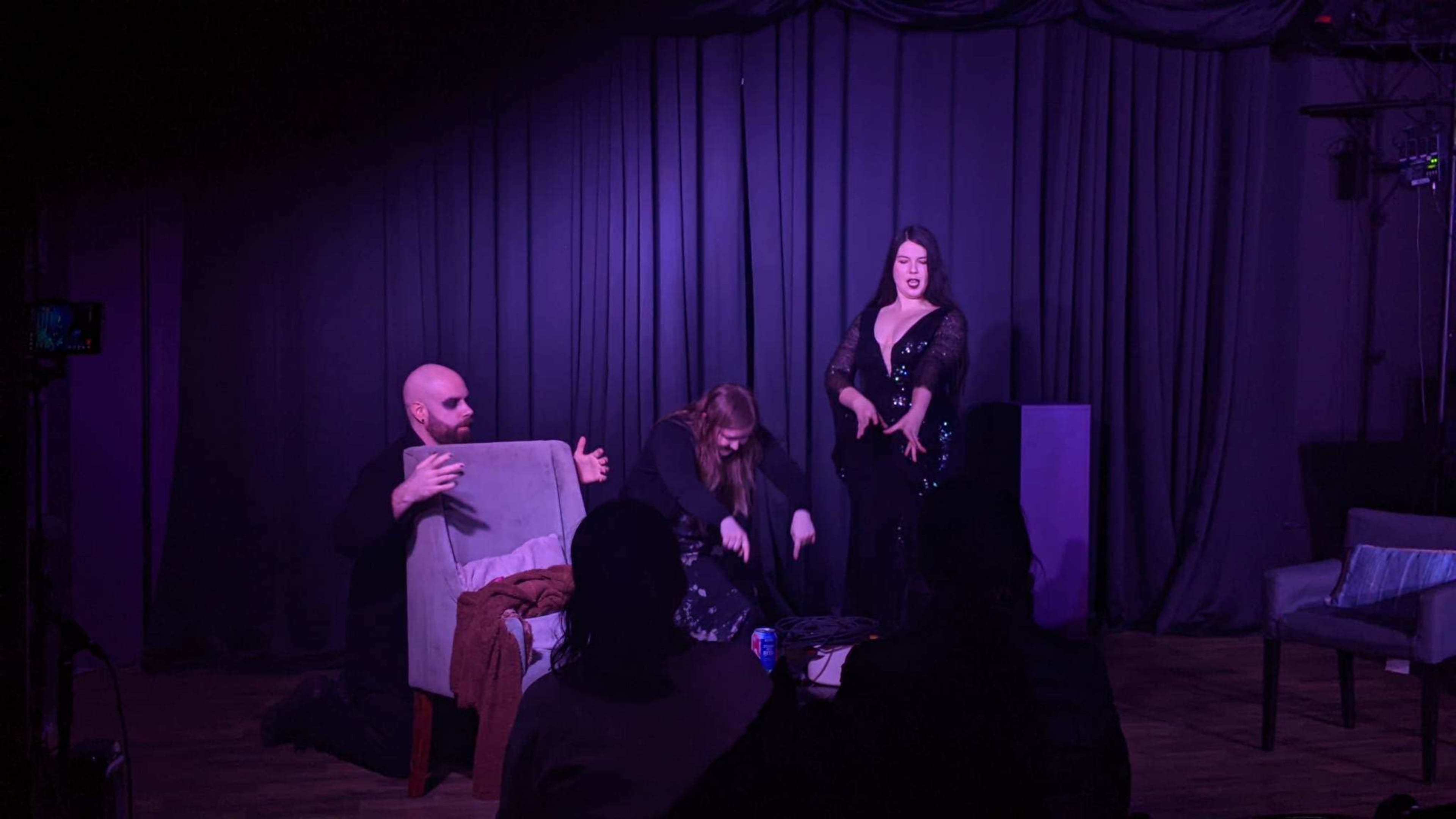 A performer in a black sequined outfit stands confidently on stage while two individuals interact with a chair in front of her.