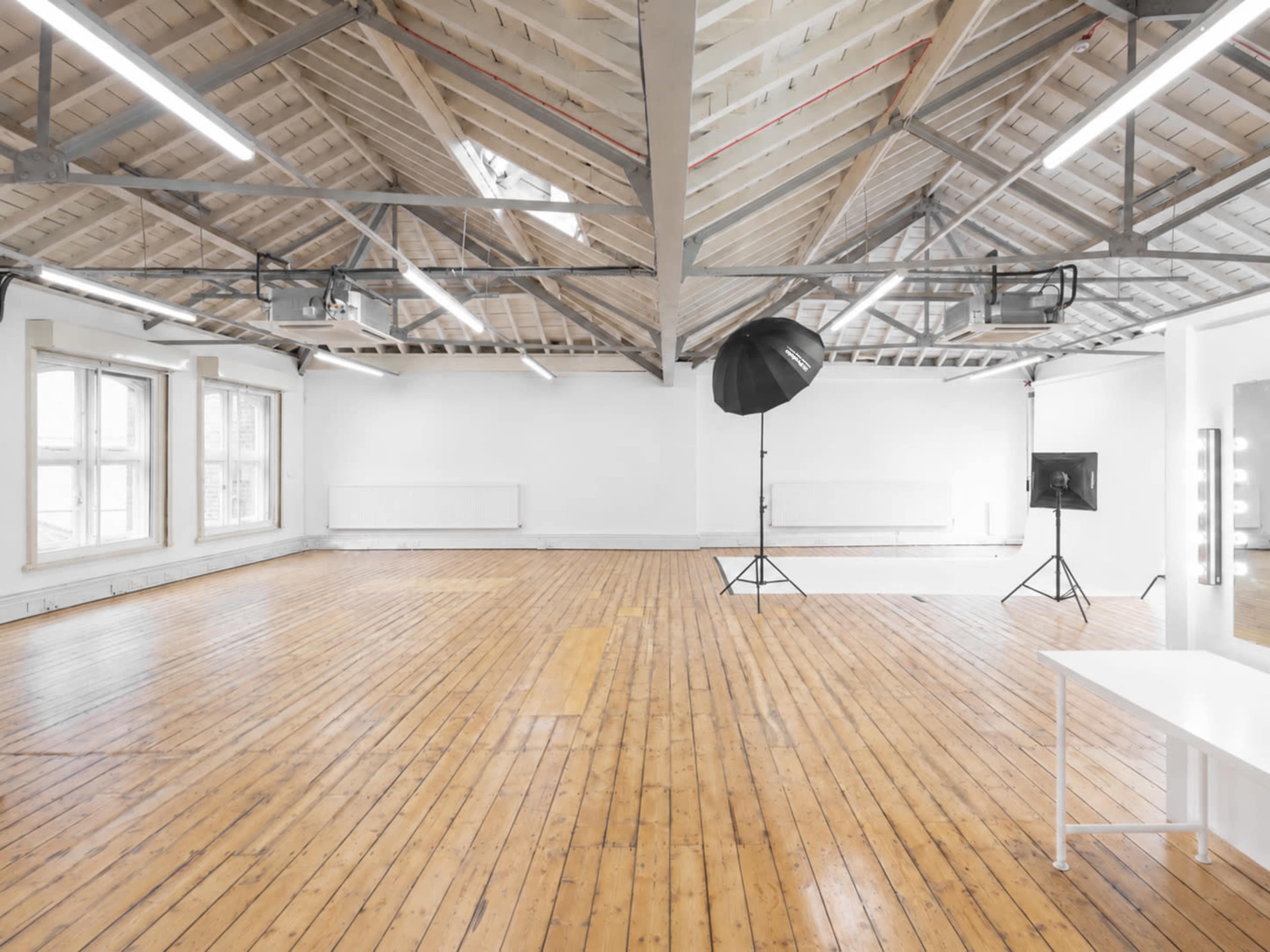 The image shows a spacious, well-lit photography studio with wooden floors, large windows, and minimalist furnishings.