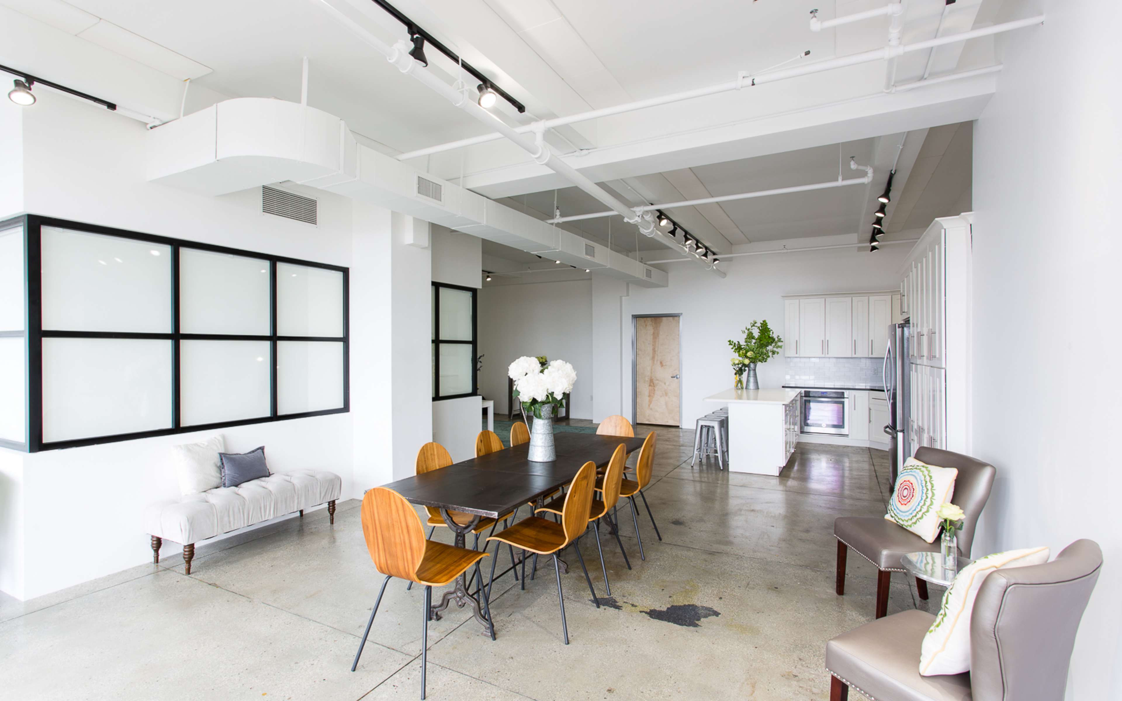 A modern, minimalist dining area featuring a long wooden table surrounded by wooden chairs, with a small kitchen in the background and large windows letting in natural light.
