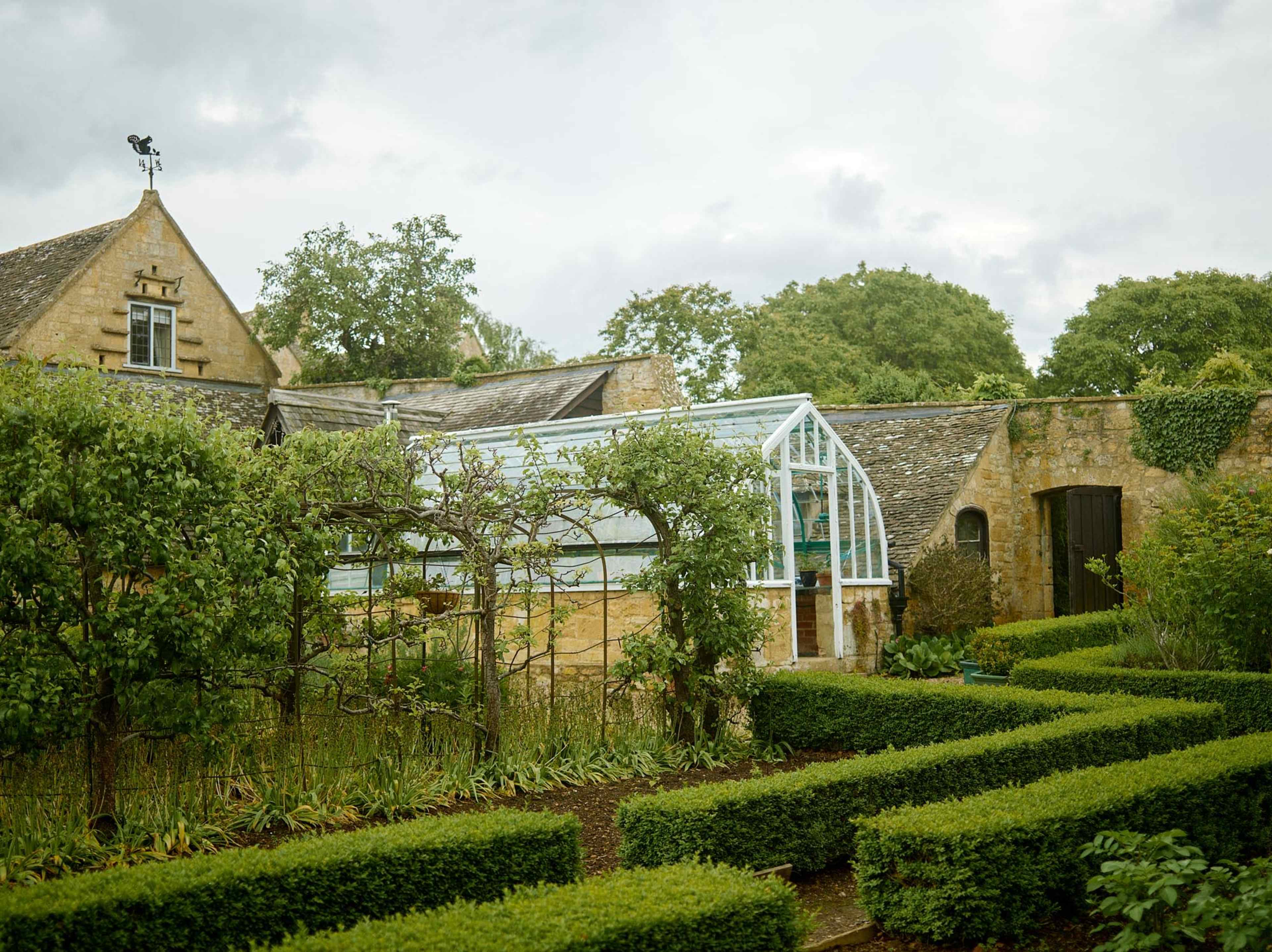 A greenhouse surrounded by neatly trimmed hedges and a stone house in a lush garden setting.