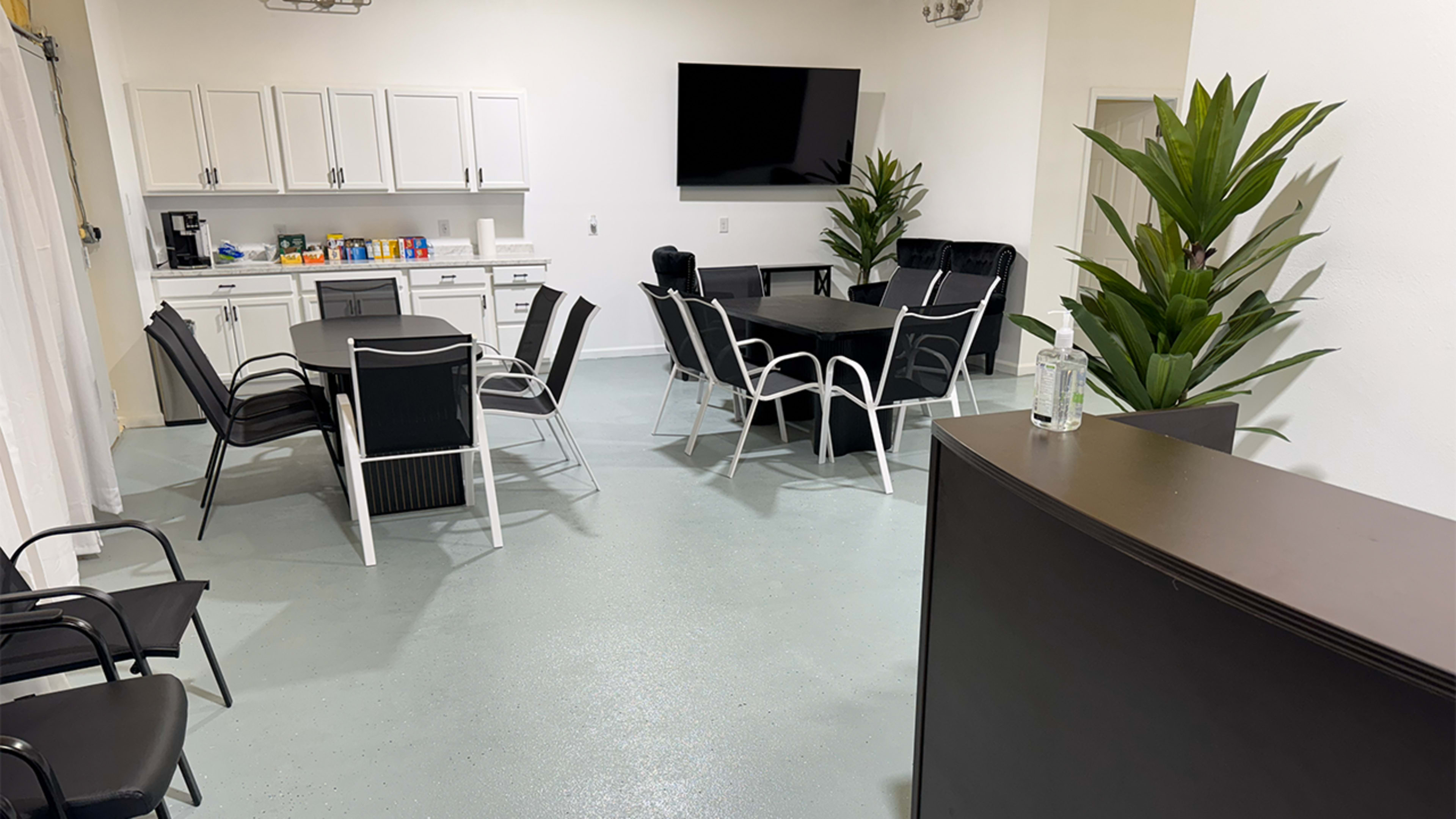 The image shows a modern conference room with a large table surrounded by chairs, a kitchenette area, and plants in the corners.