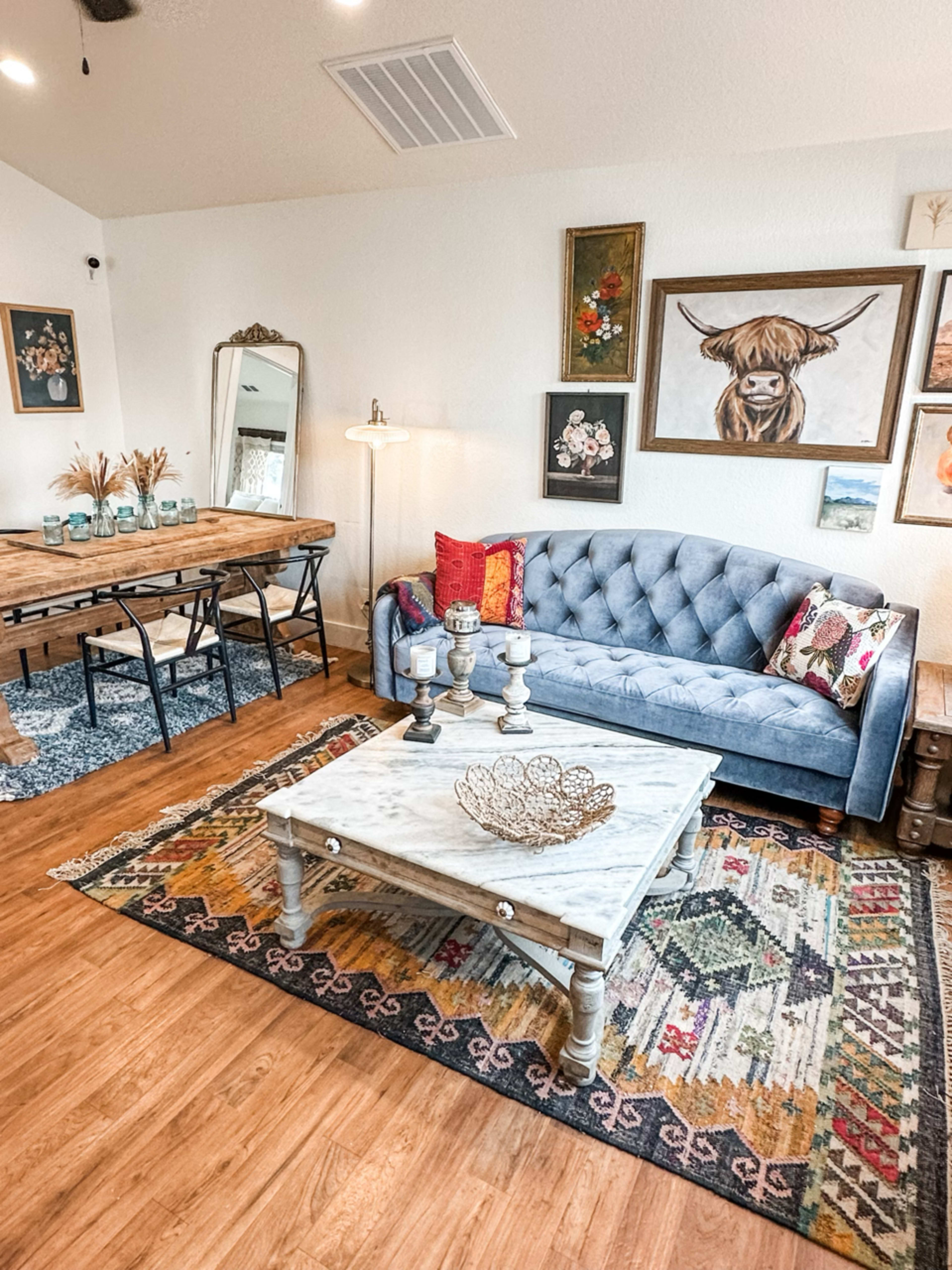 A cozy living room with a blue couch, a wooden dining table, a patterned area rug, and various wall decorations including animal art.