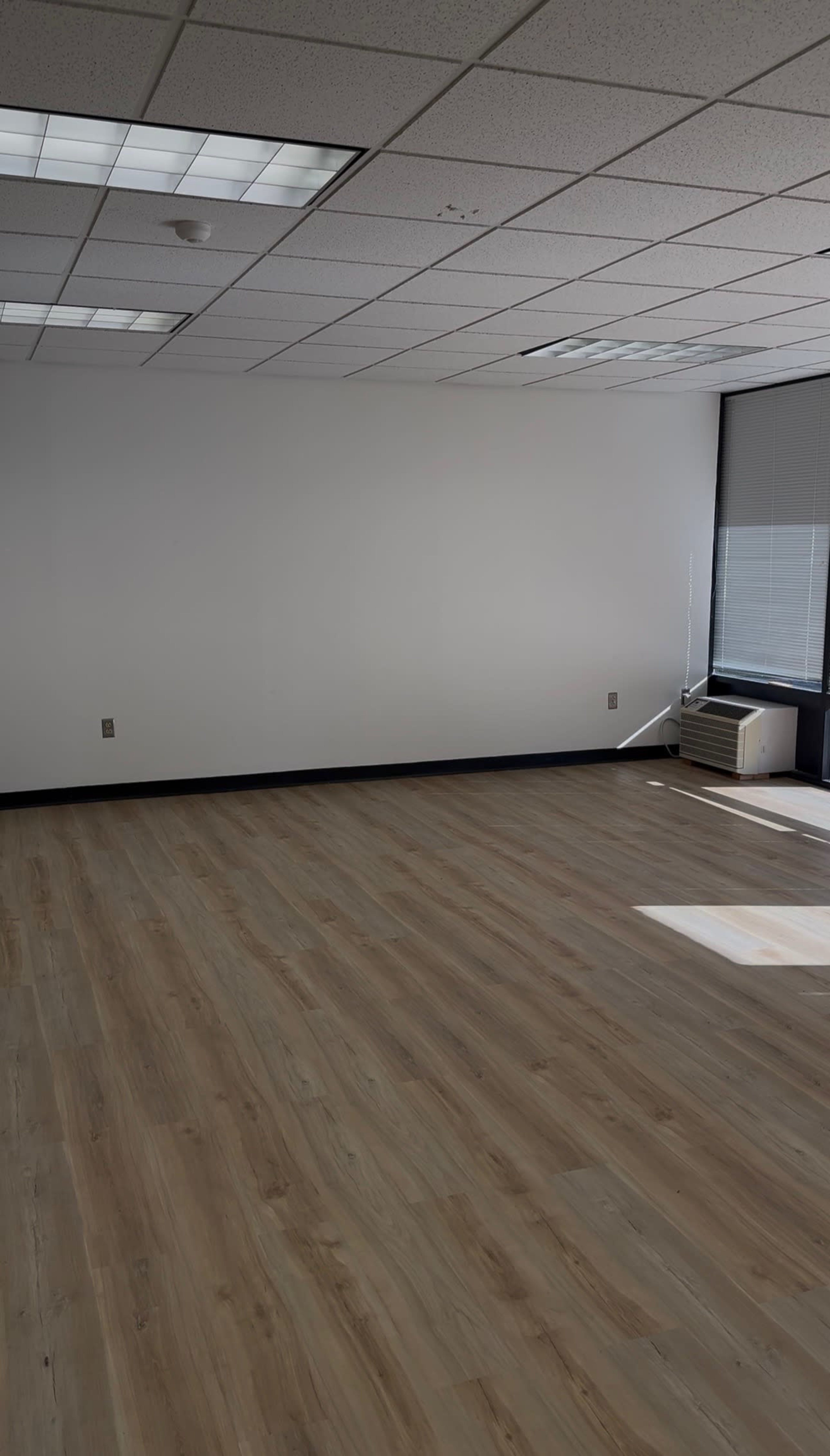 The image shows an empty office space with a wooden floor and a single window letting in natural light.