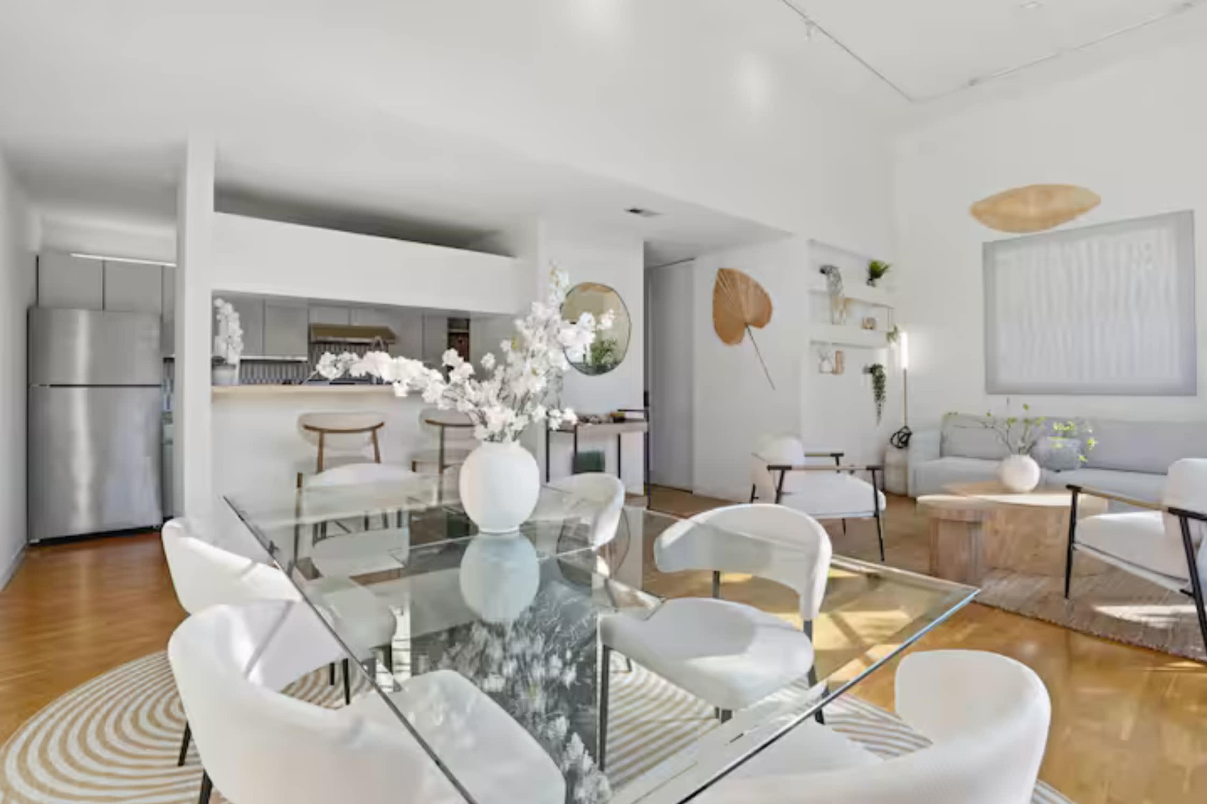 The image shows a bright, modern dining area with a glass table surrounded by white chairs, leading to an open kitchen space.