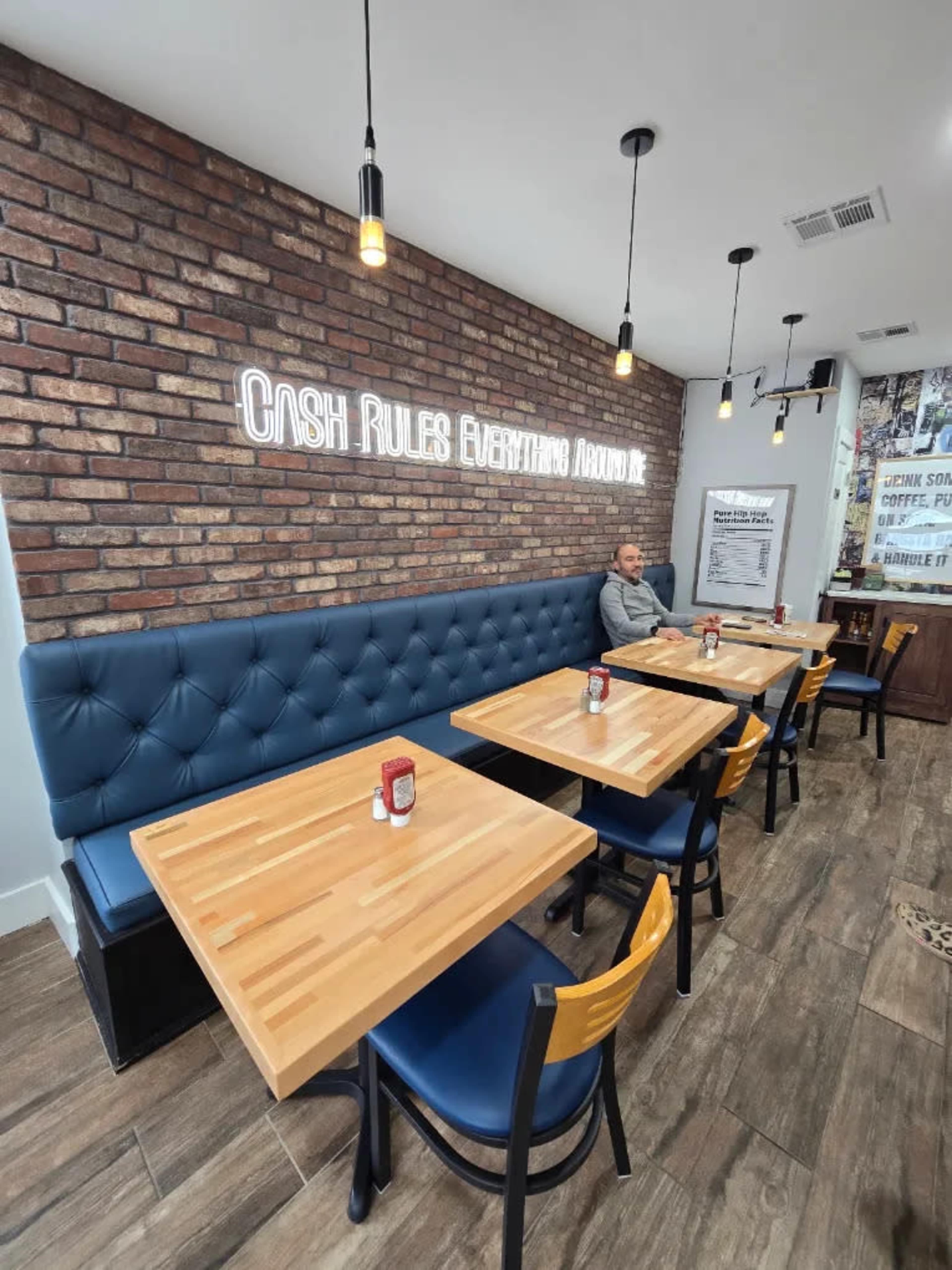The image features a modern café interior with wooden tables, blue leather seating, and a brick wall displaying the phrase "CASH RULES EVERYTHING AROUND ME."