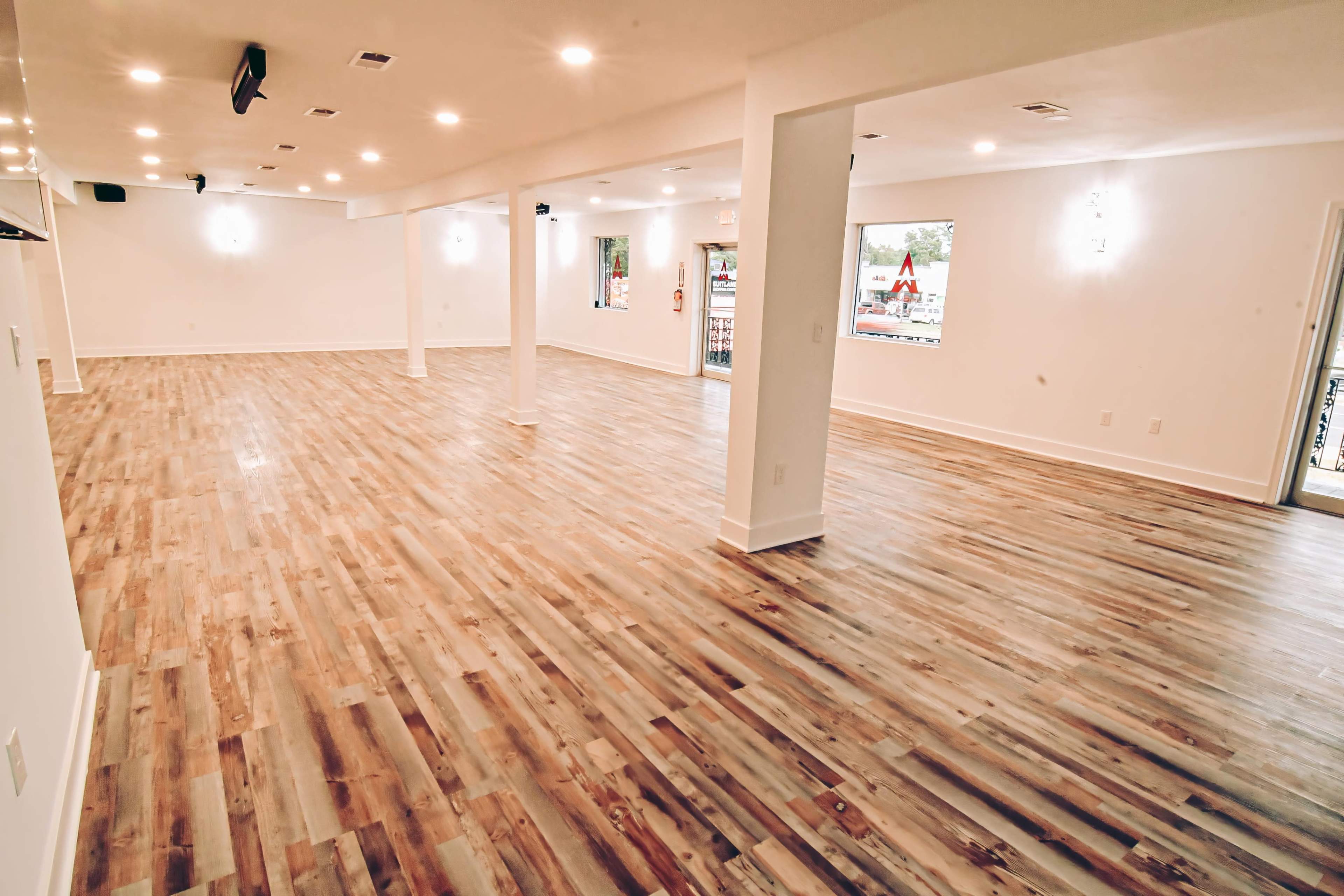 The image shows a spacious, empty room with wooden flooring and white walls, illuminated by overhead lights.